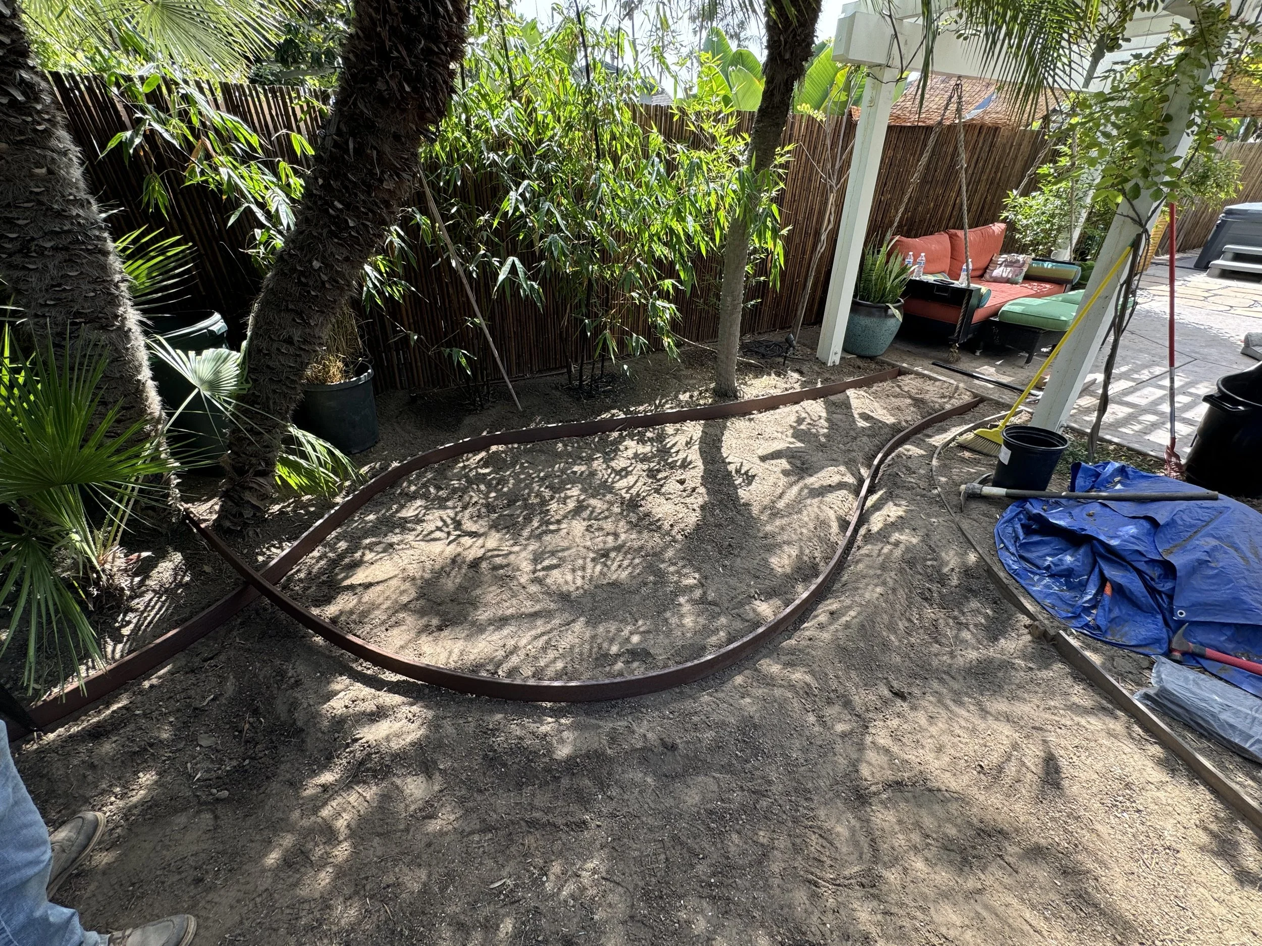 A backyard area under renovation with a curved retaining wall frame on the ground, surrounded by trees and potted plants. There is a outdoor couch with cushions and a glass table, along with construction tools and materials including a tarp, buckets, and a broom.