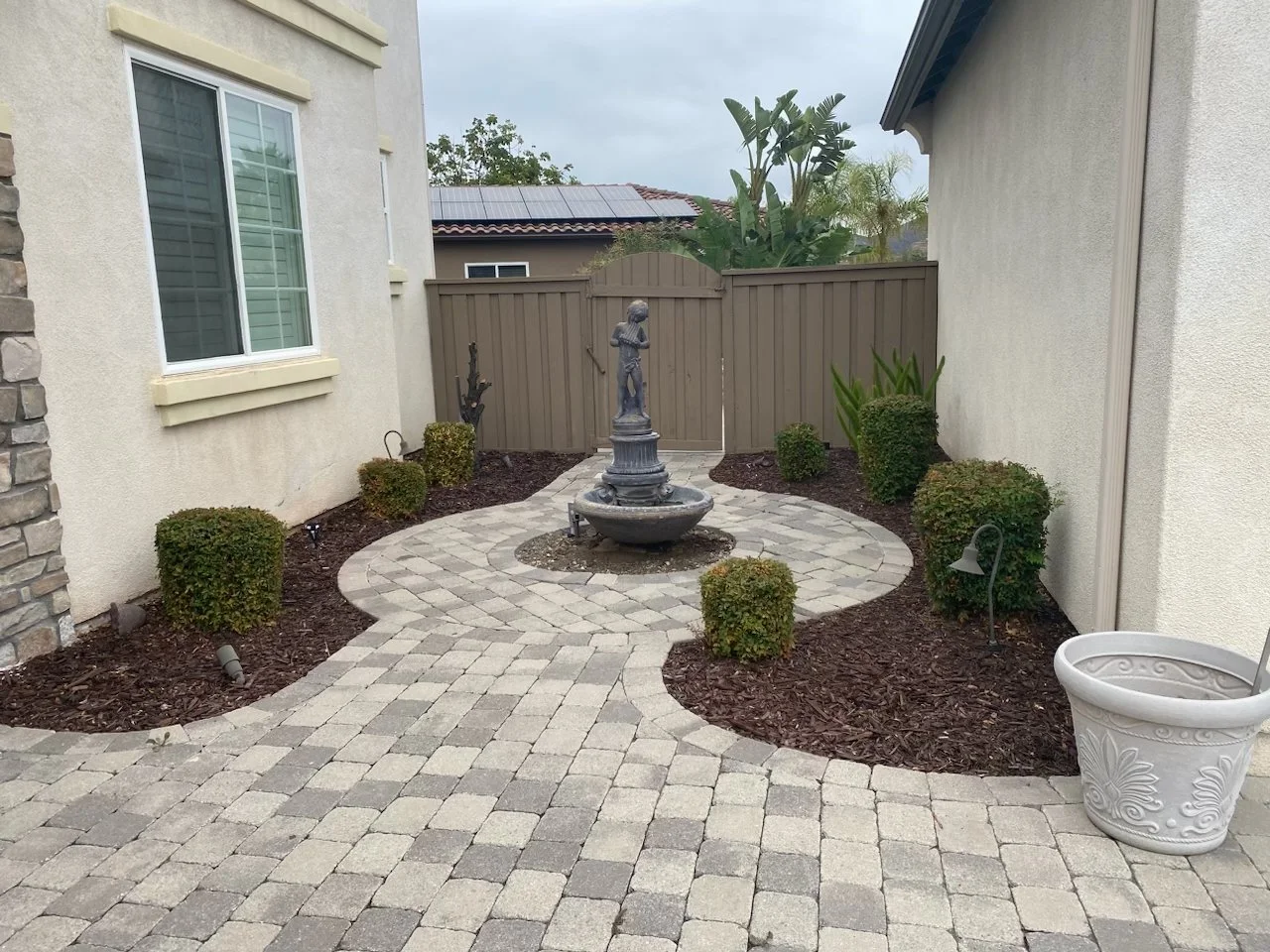 Small backyard with a stone pathway, a central fountain with a sculpture, surrounded by small bushes, a fence, and potted plants.