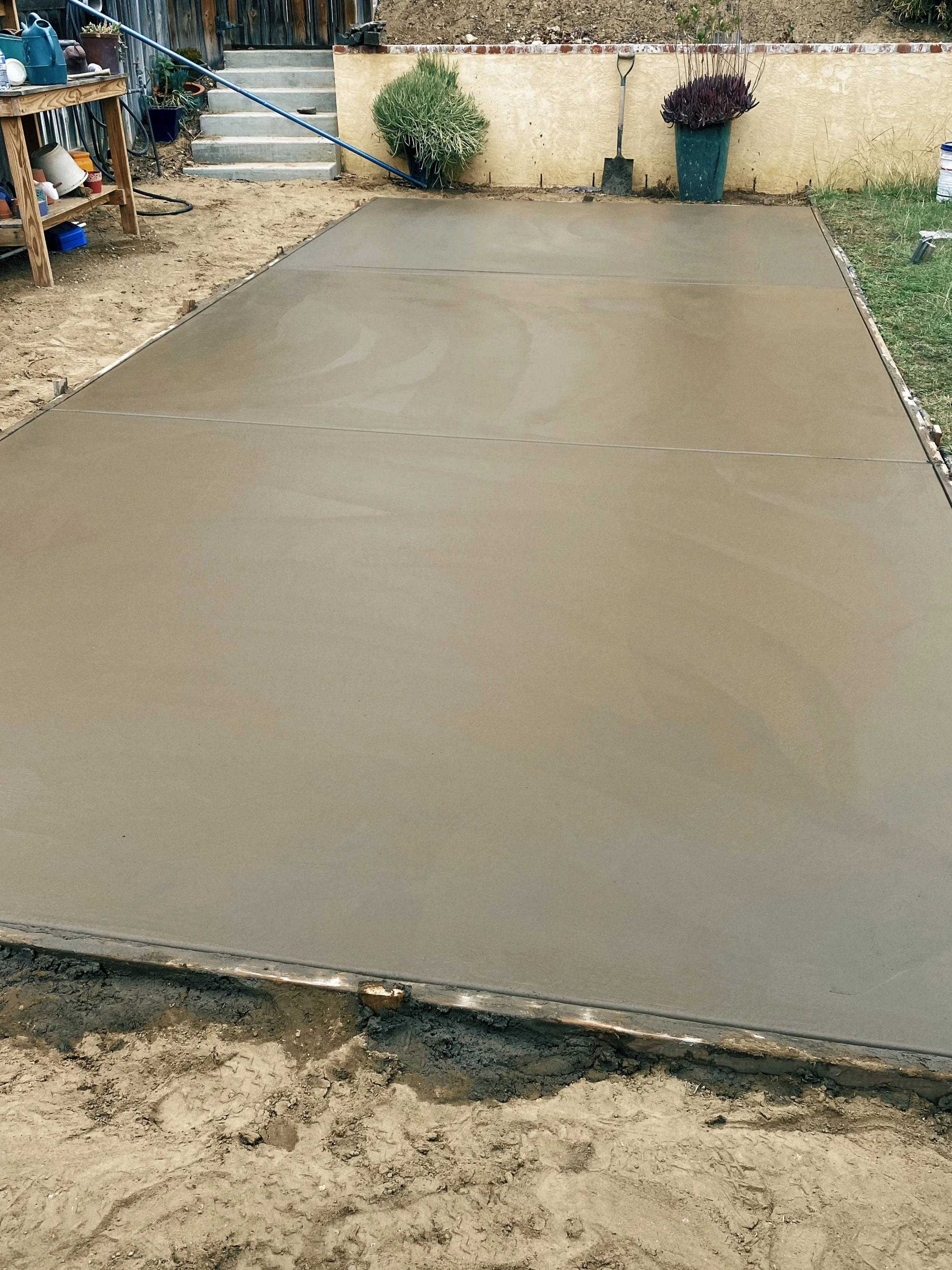 Freshly poured concrete slab in a backyard, with garden tools and plants in the background.