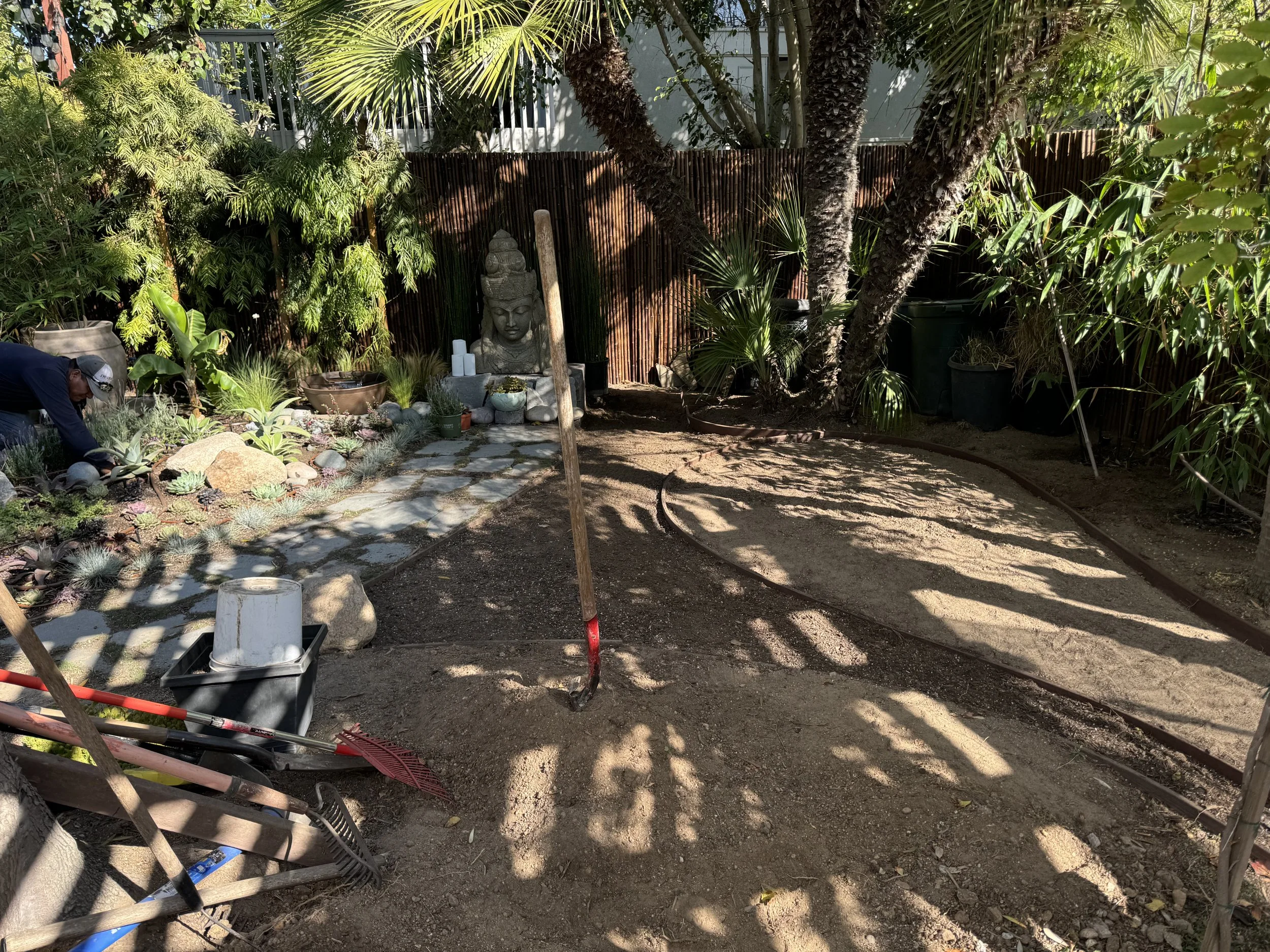 Garden with a stone pathway, trees, plants, and a Buddha statue in the background, with tools and landscaping materials nearby.
