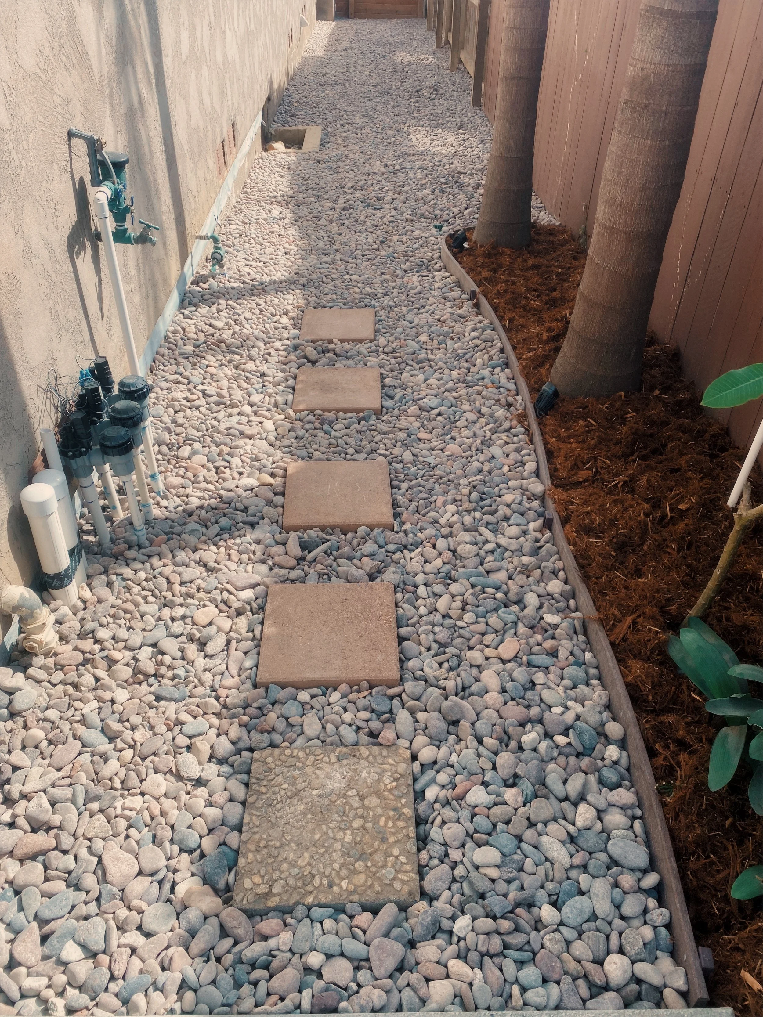 A backyard pathway made of square stepping stones surrounded by small pebbles, with a wooden fence on the right side and a house wall on the left side, with trees planted along the fence.