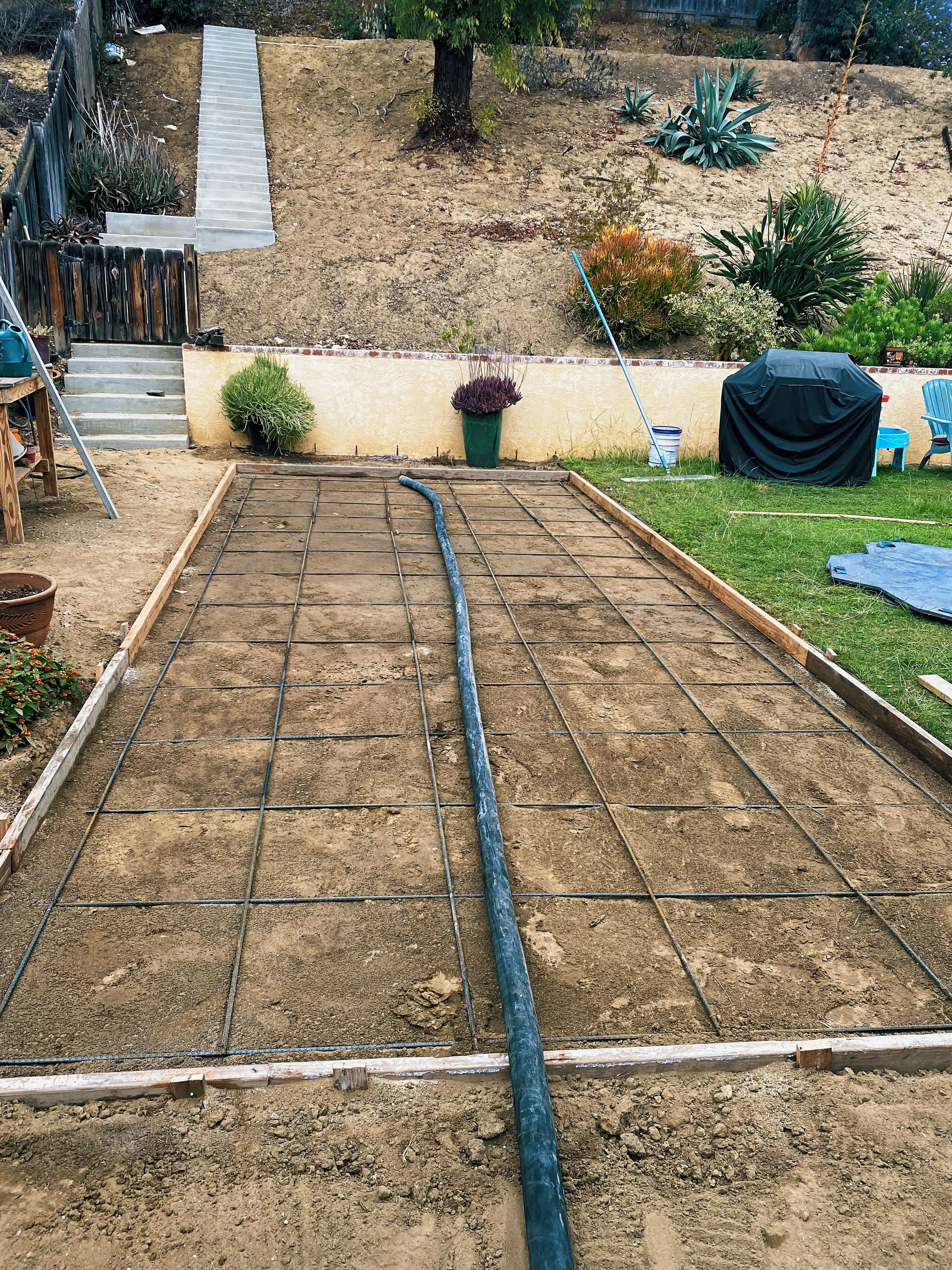 A backyard with a dirt area framed with wooden boards, a black hose running through it, and a green lawn with a covered grill, a blue chair, and gardening tools. In the background, there are stairs leading up a slope with plants and trees.