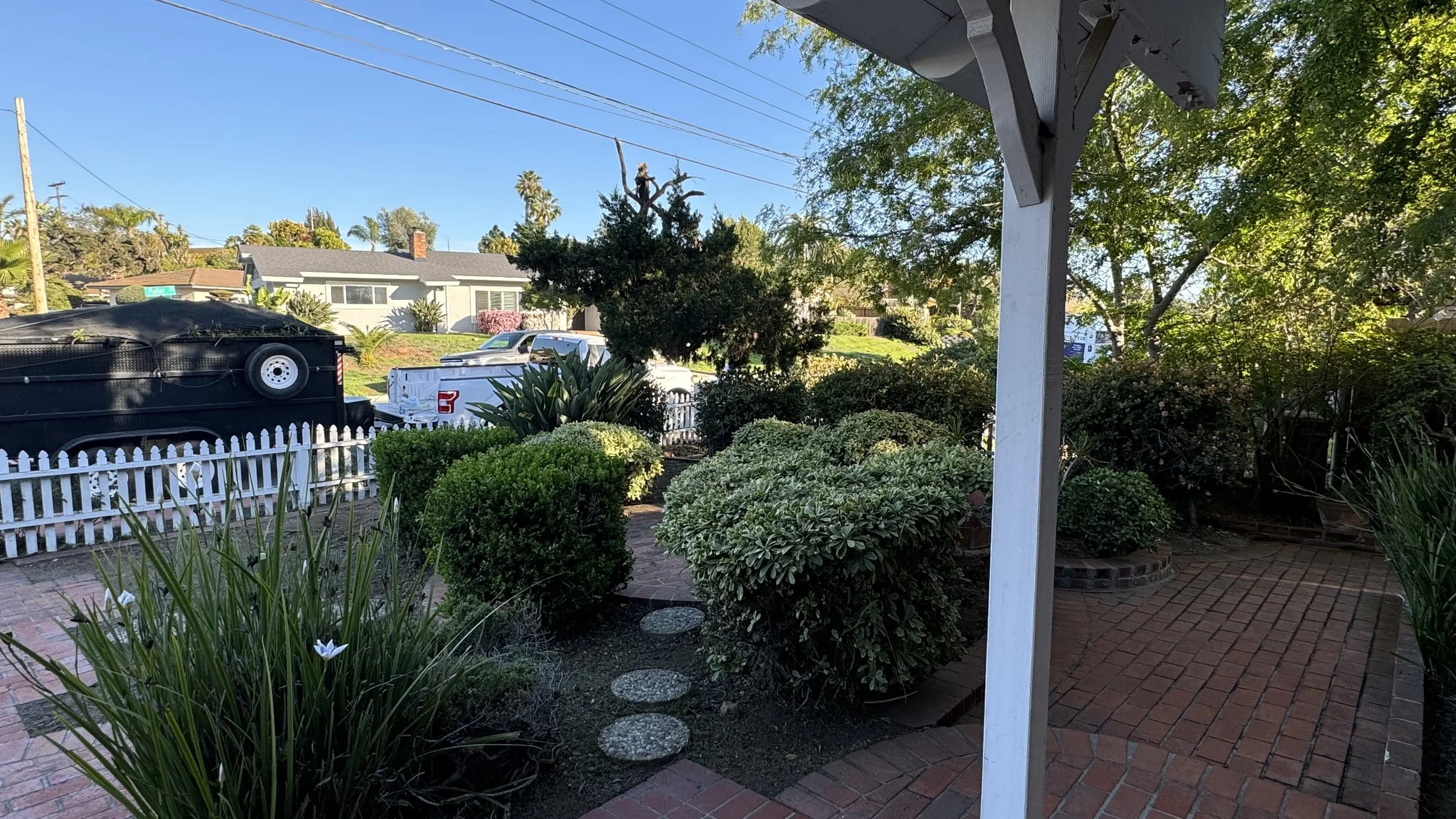A front yard with a brick pathway, various green bushes, and trees, with a white picket fence, and a black trailer and white truck parked on the street in the background.