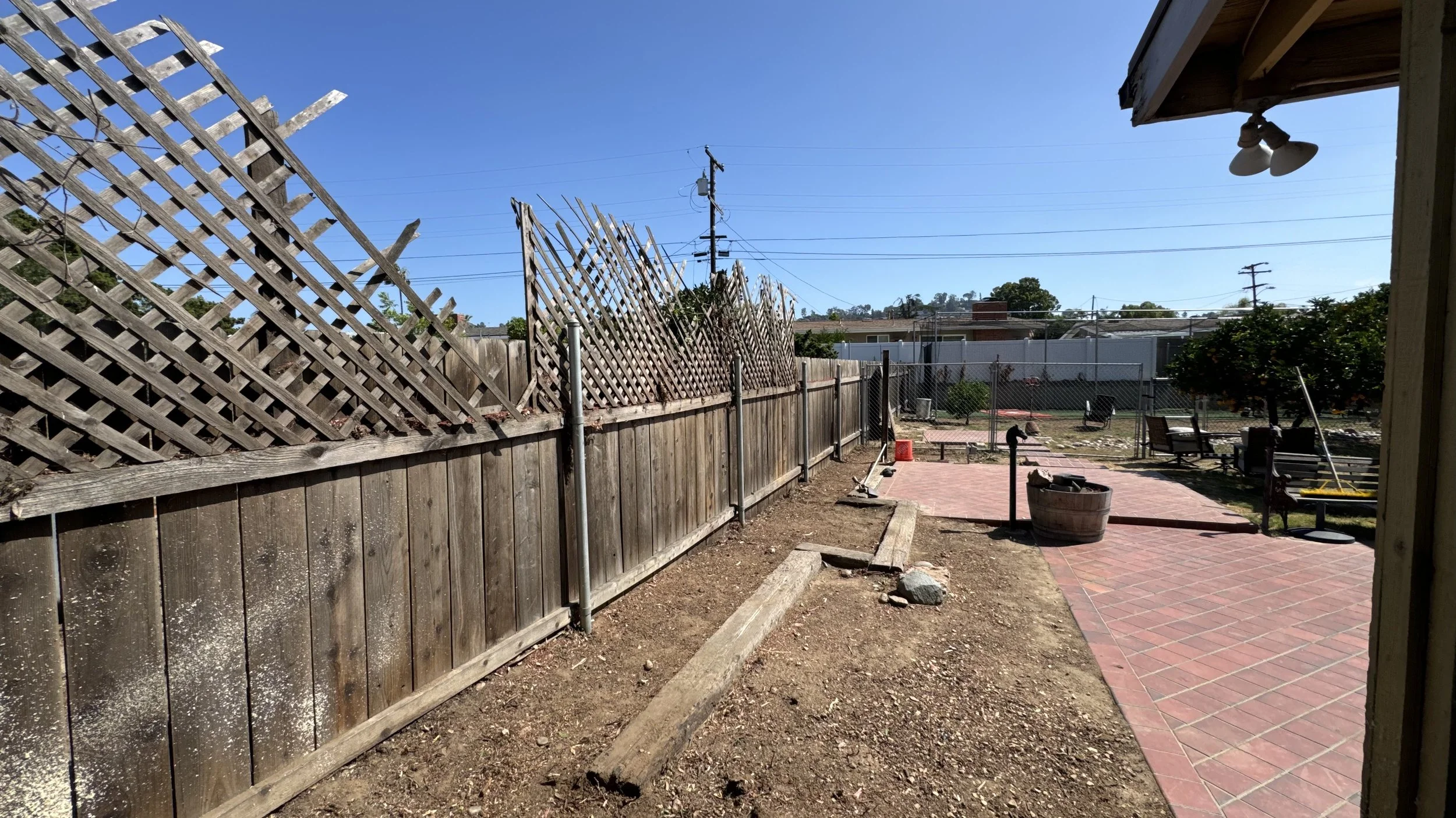 Partially renovated backyard with a wooden fence, some sections of lattice torn down, a paved patio area with benches and a small garden, and a clear blue sky.