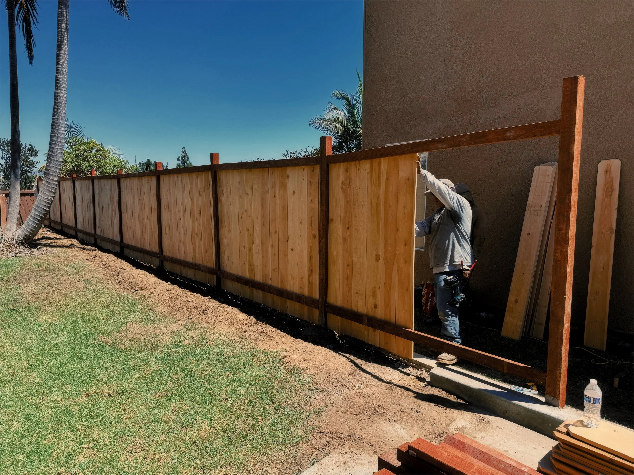 Person installing a wooden fence near a house, with a water bottle and building materials nearby.