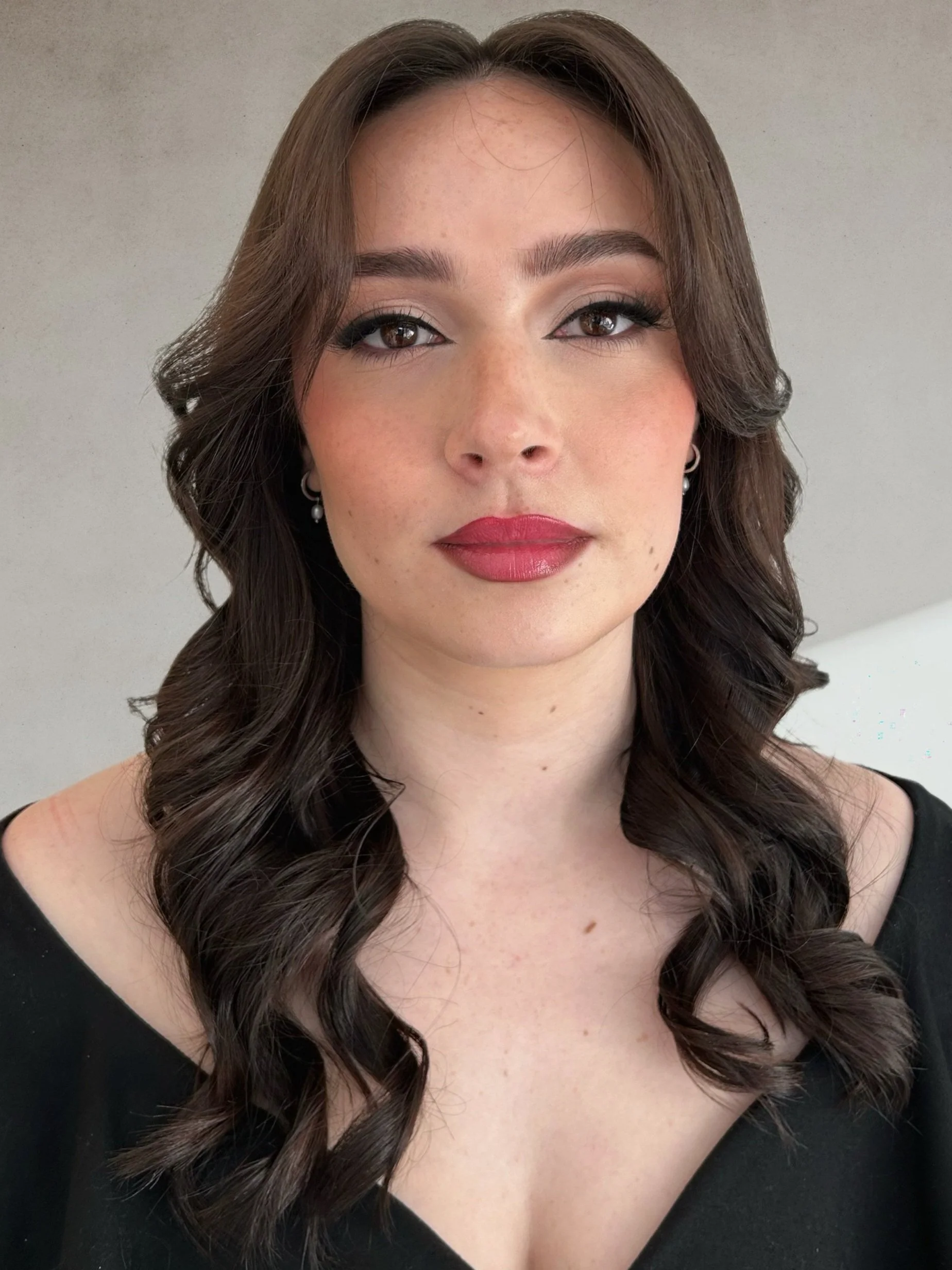 A portrait of a woman with wavy brown hair, makeup with red lipstick, earrings, and wearing a black top.