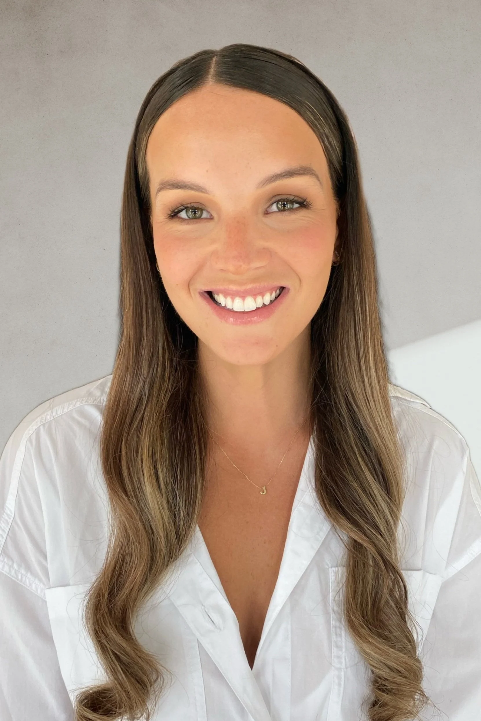 A woman with long brown hair, light skin, green eyes, and a bright smile, wearing a white shirt and a delicate gold necklace with a small pendant, against a plain gray background.