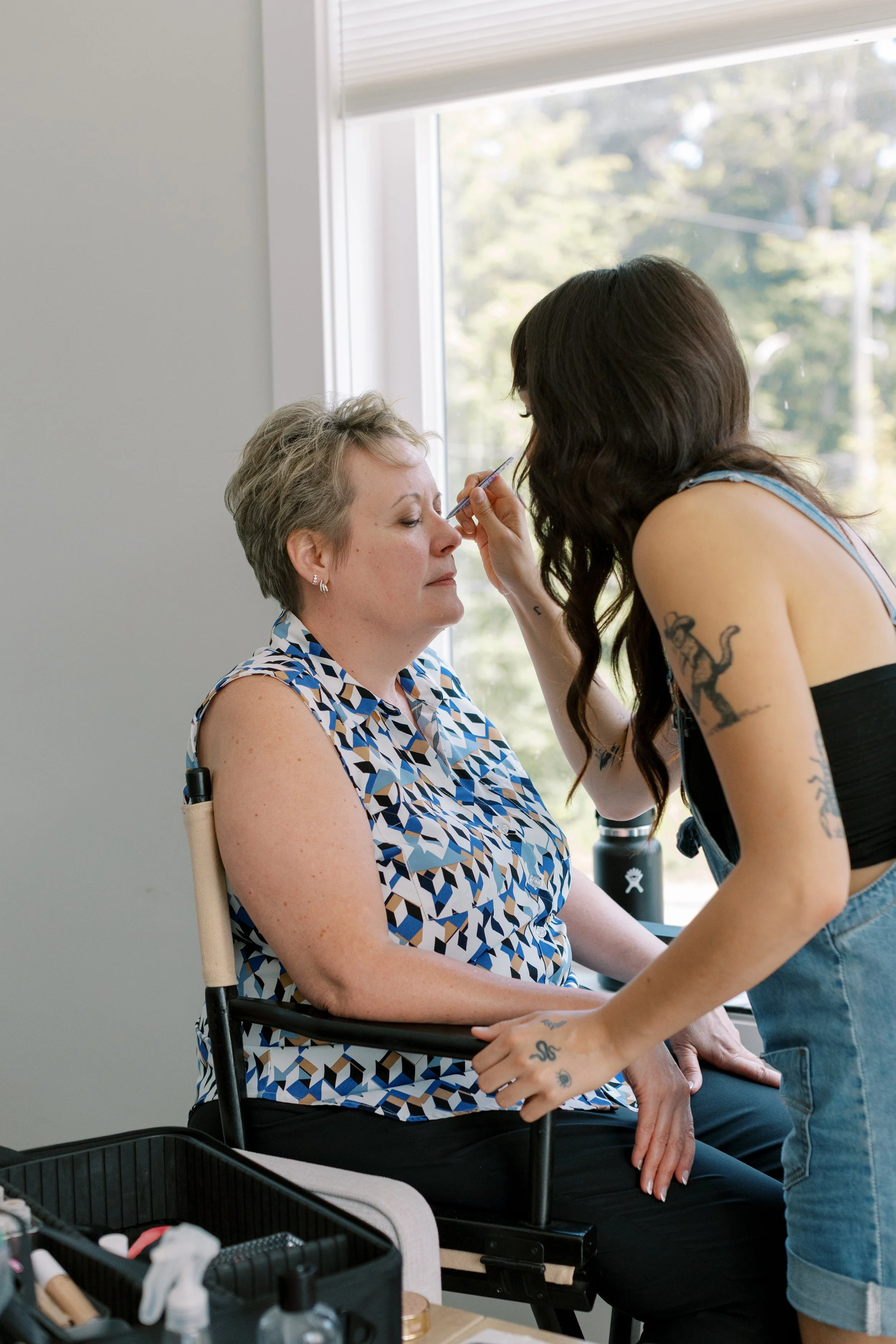 Woman in a patterned sleeveless blouse having makeup applied by a makeup artist with tattoos, sitting near a window with trees outside.