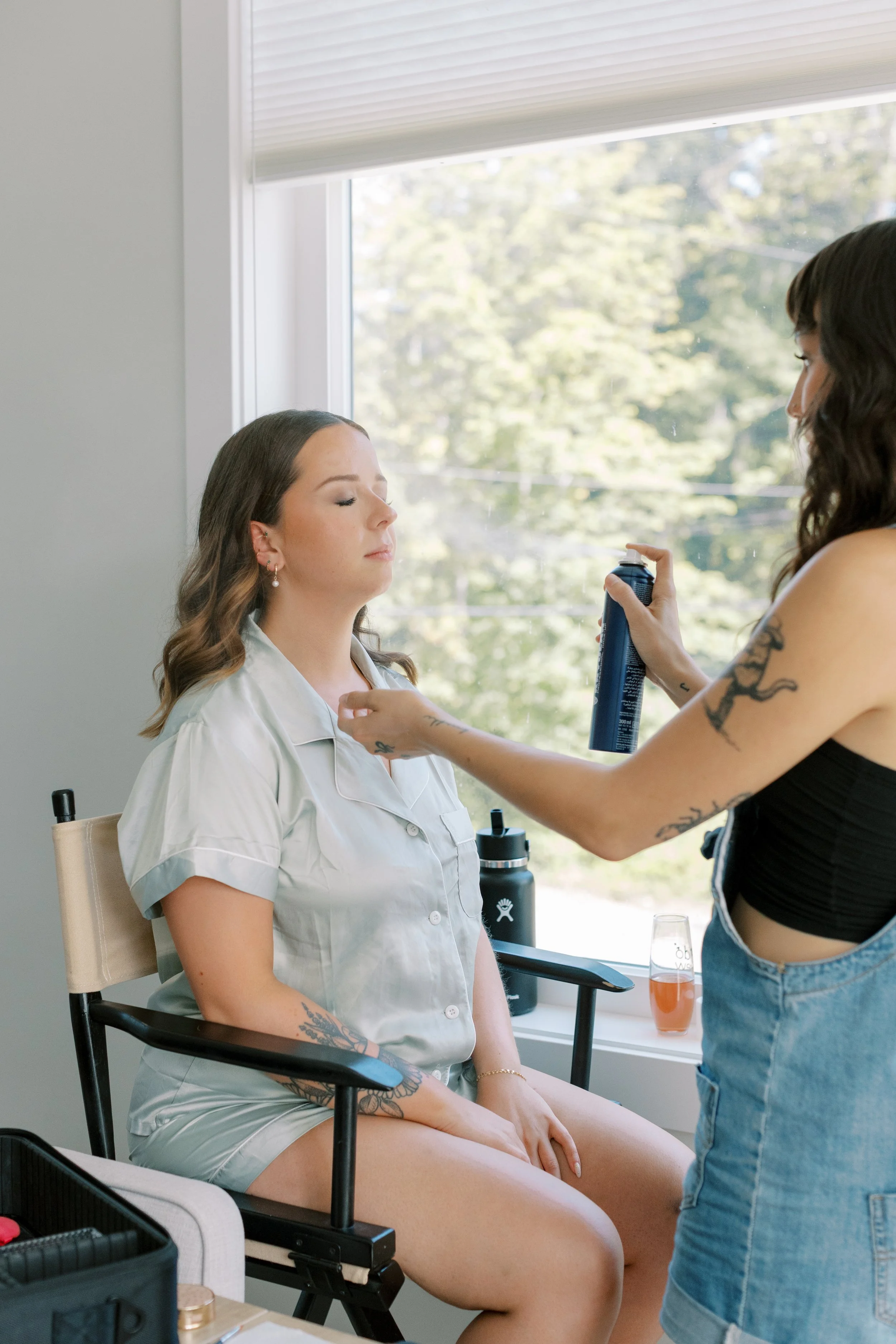 A woman sitting with her eyes closed while another woman sprays something on her face in a bright room with a window showing trees outside.