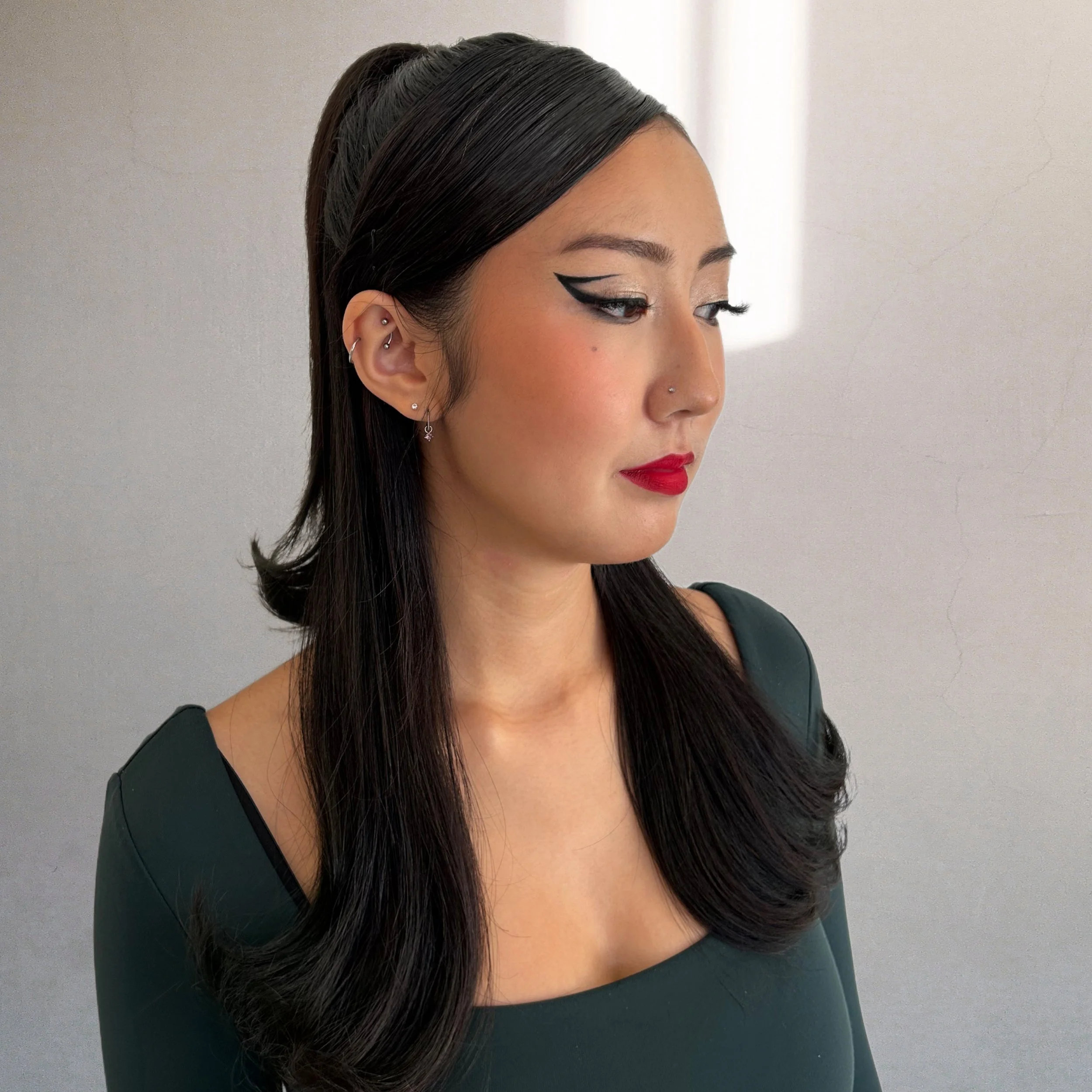 A woman with long dark hair styled in loose curls, wearing bold makeup with winged eyeliner and red lipstick, looking to the side, against a plain background.