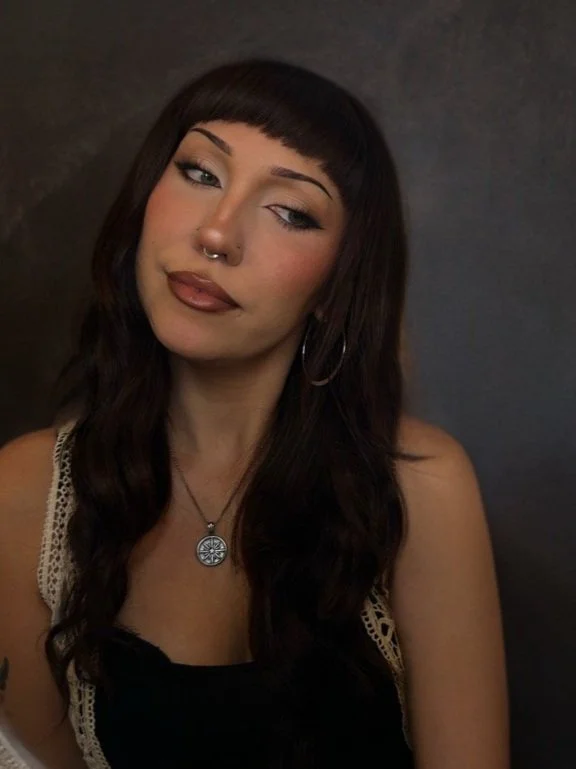 A young woman with dark brown hair and bangs, wearing hoop earrings, a nose ring, and a pendant necklace, posing in front of a dark background.