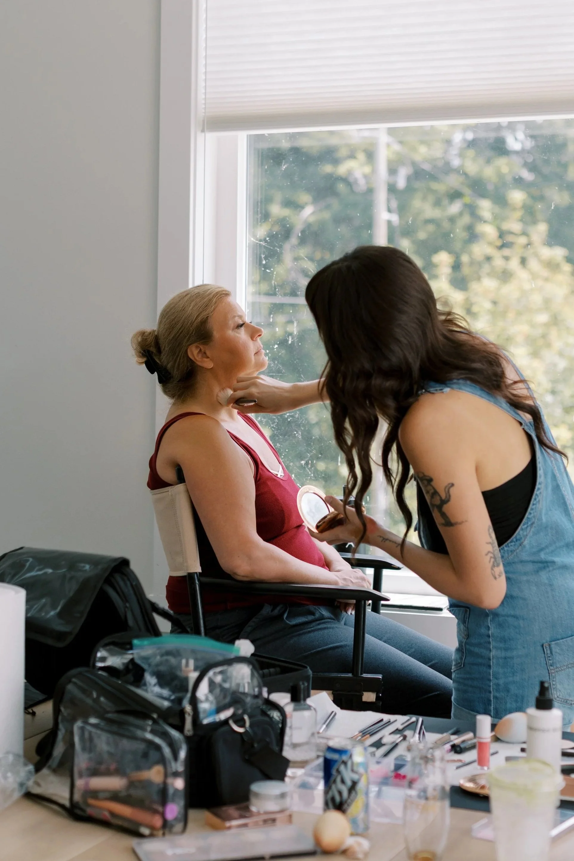 A makeup artist applying makeup to a woman sitting near a window, with makeup tools and supplies on a table in front of them.