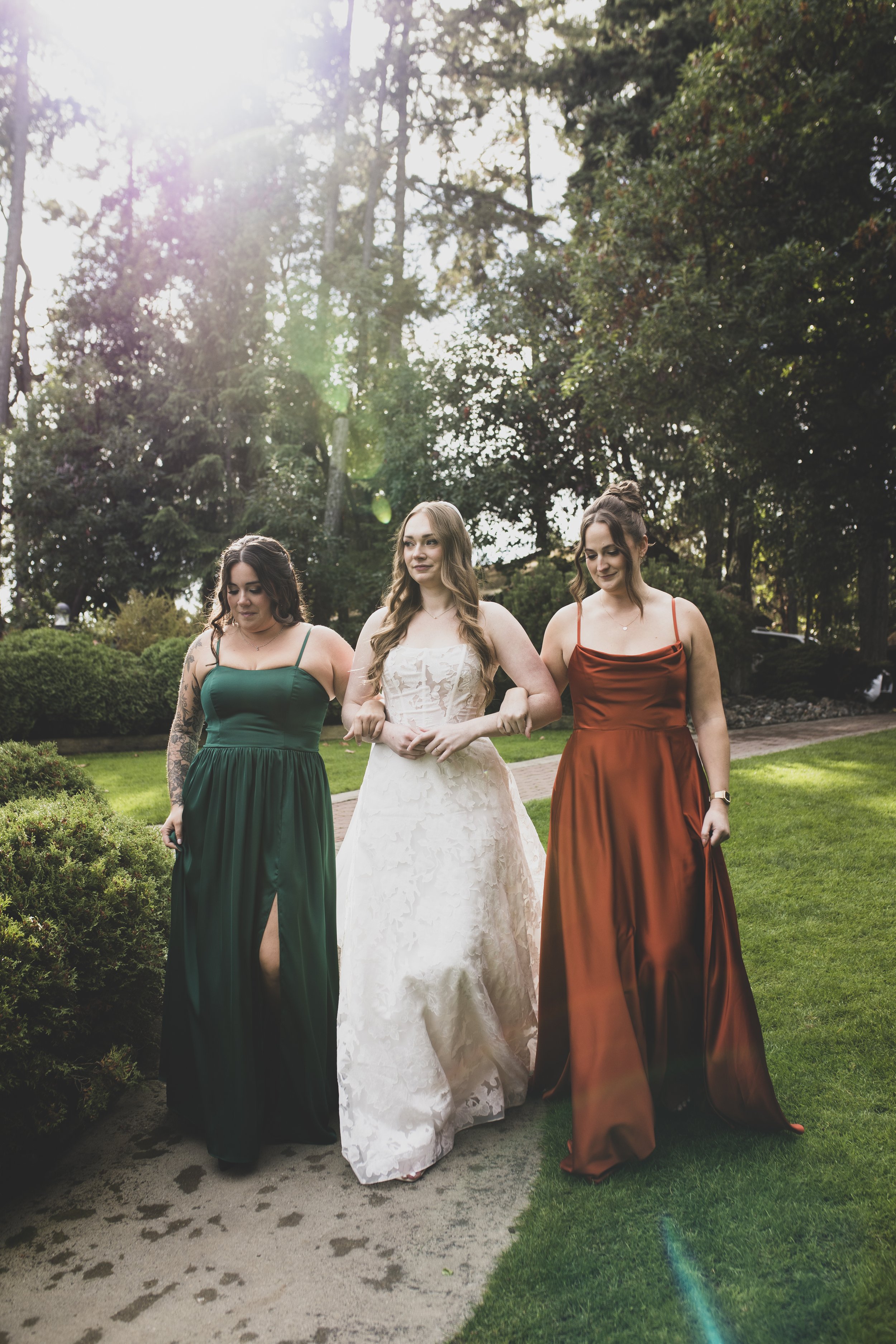 Three women in elegant dresses walking arm-in-arm outdoors on a garden path, with sunlight filtering through trees in the background.