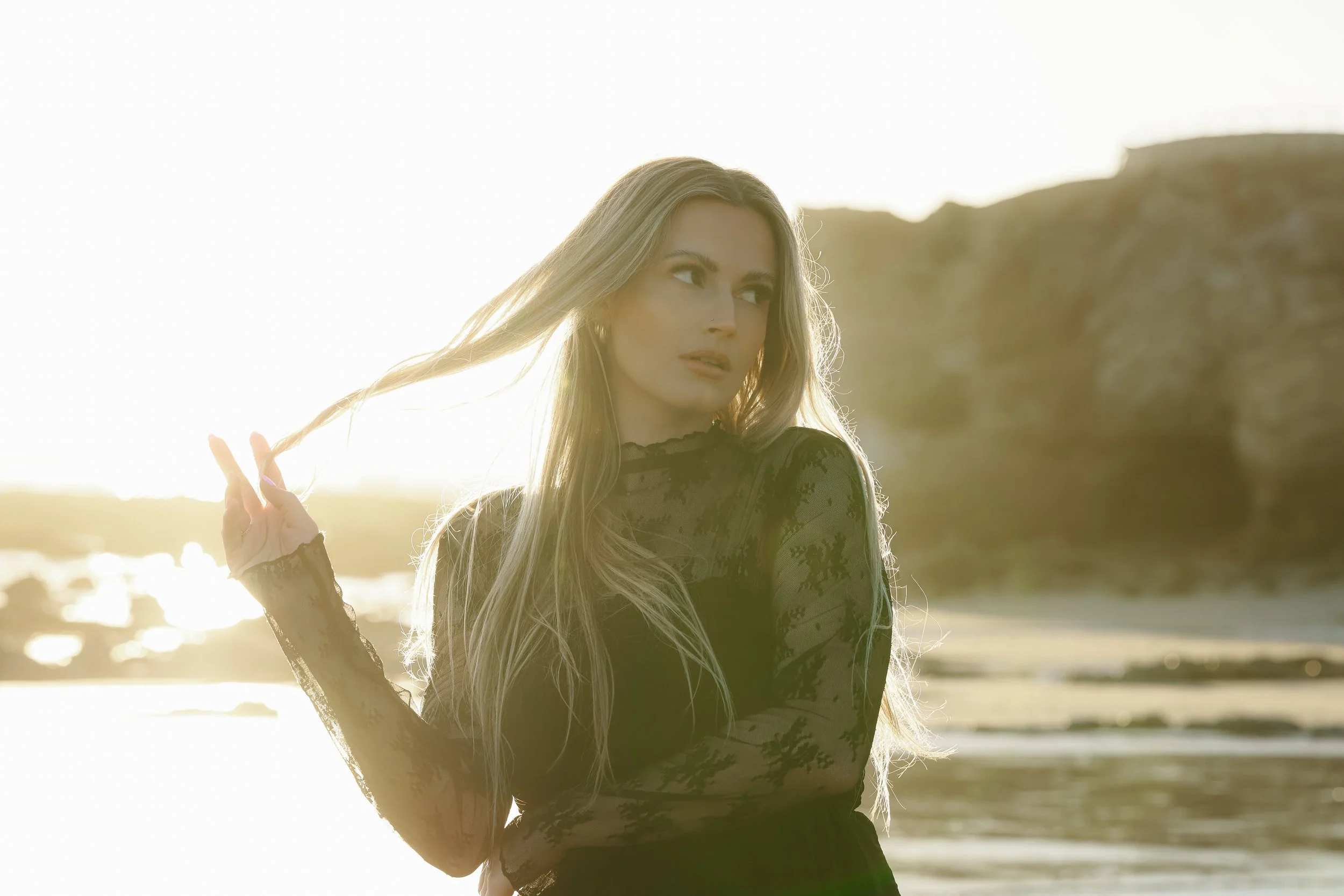 A young woman with long blonde hair stands outdoors near a railing and a large bush, dressed in a black long-sleeve shirt and blue jeans, with the setting sun behind her.