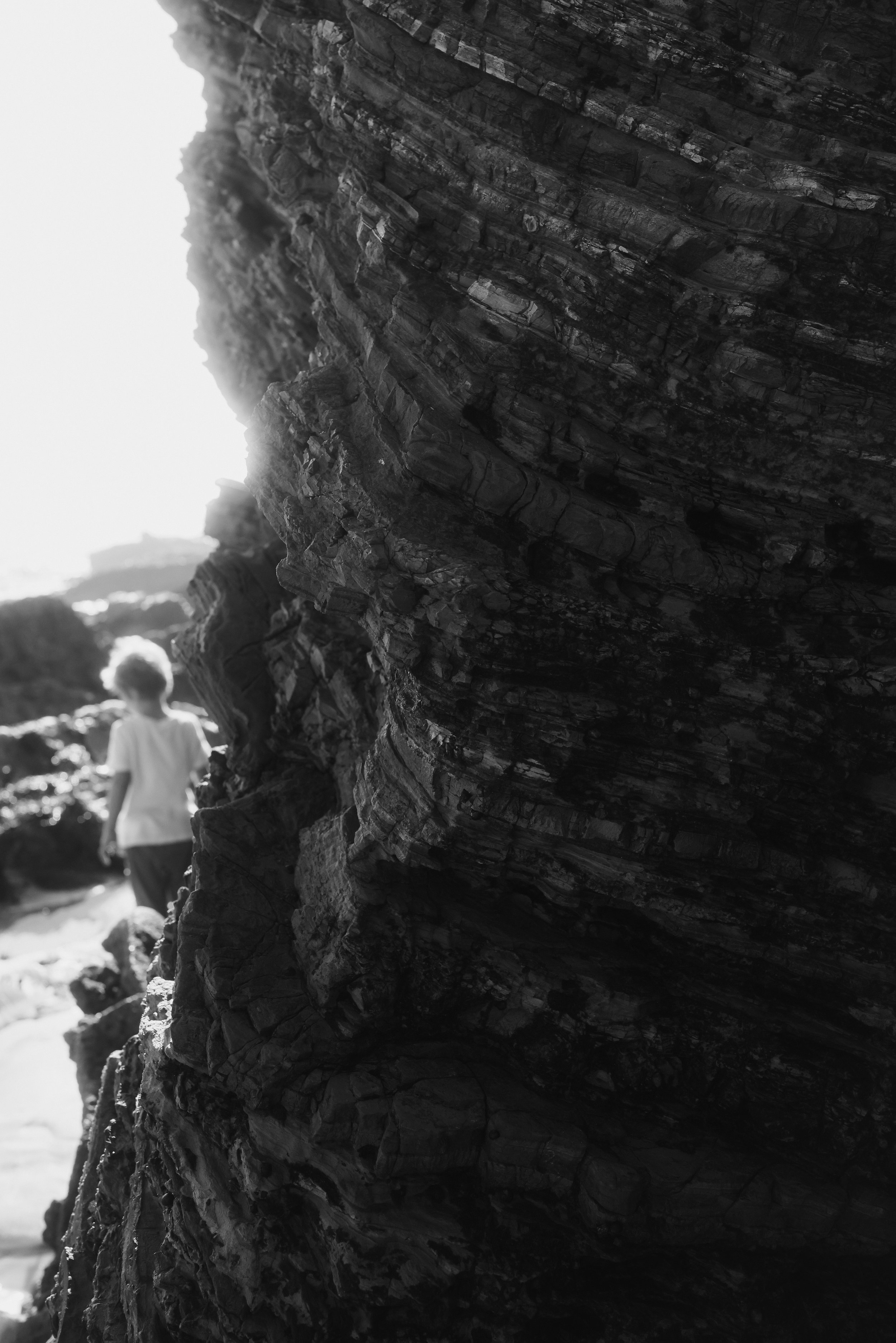 A person with light-colored hair walking along a rocky coastline with towering layered rocks, captured in black and white.