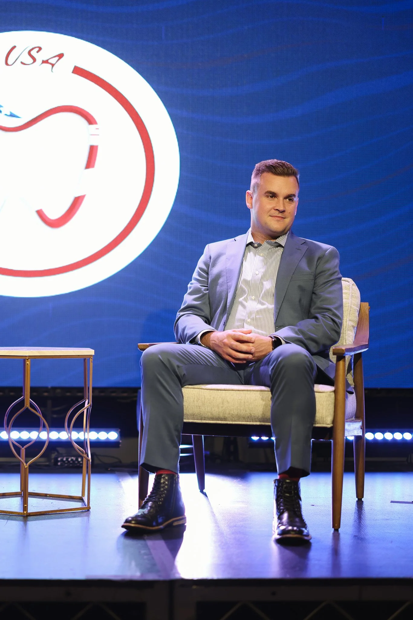 A man in a gray suit and striped shirt sitting in a chair on a stage with a large screen behind him displaying a partial logo featuring red, white, and blue colors. There is a small side table with a gold frame and glass top to his left.