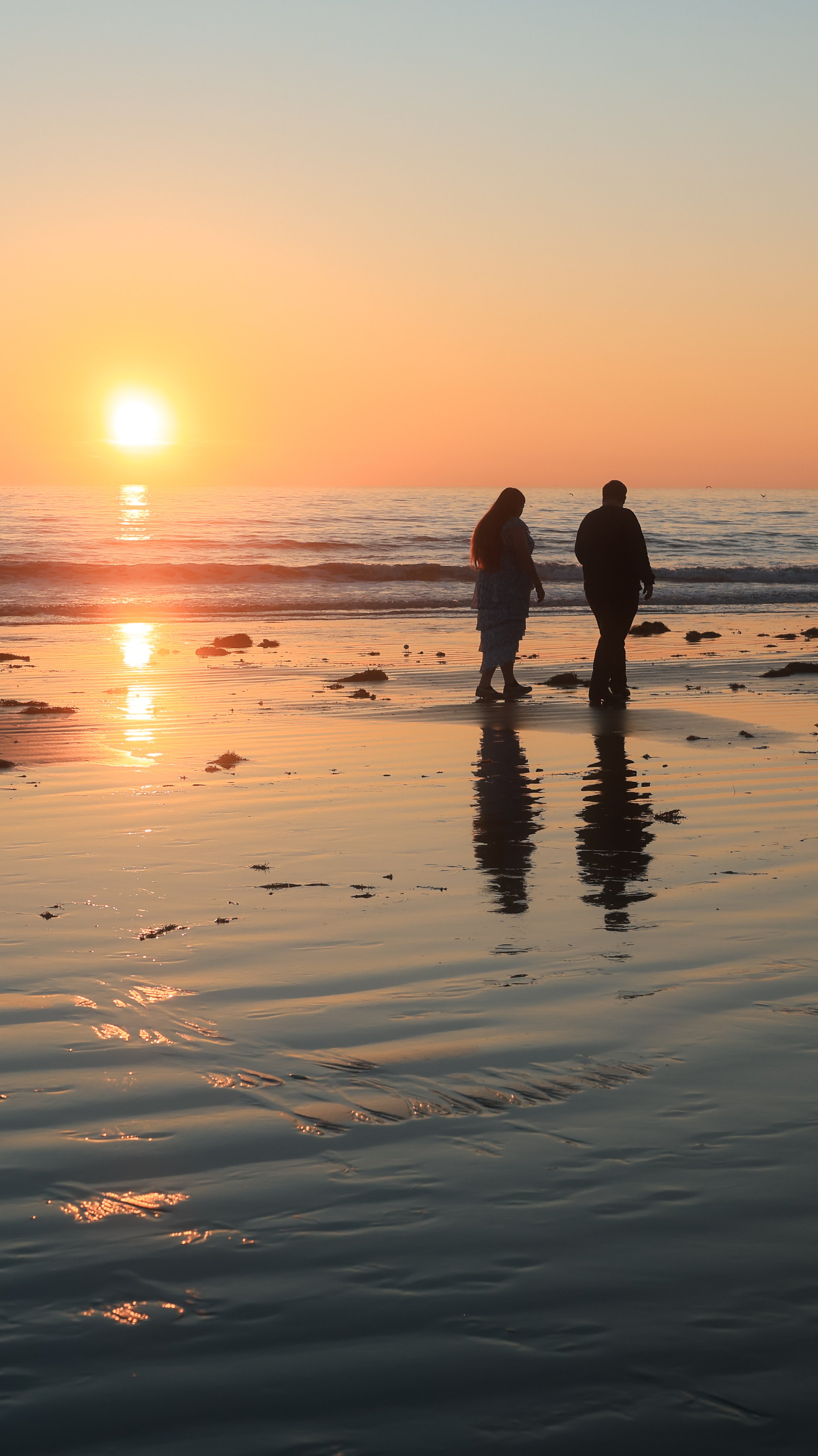 Silhouettes of a woman and a man walking on a beach during sunset with the ocean in the background.