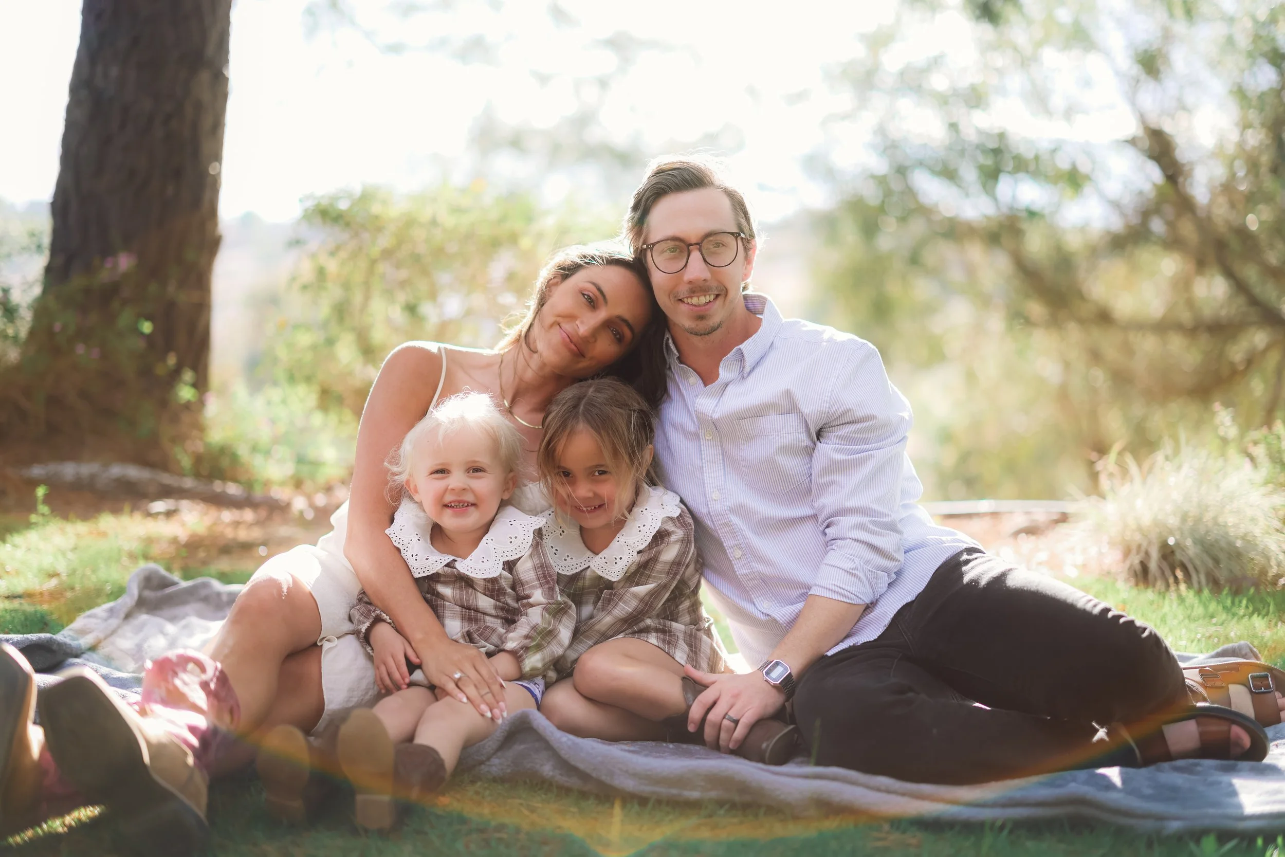 A family of four sitting on a blanket outdoors in a park, with a tree in the background. The mother, father, and two young girls are smiling and enjoying a sunny day.