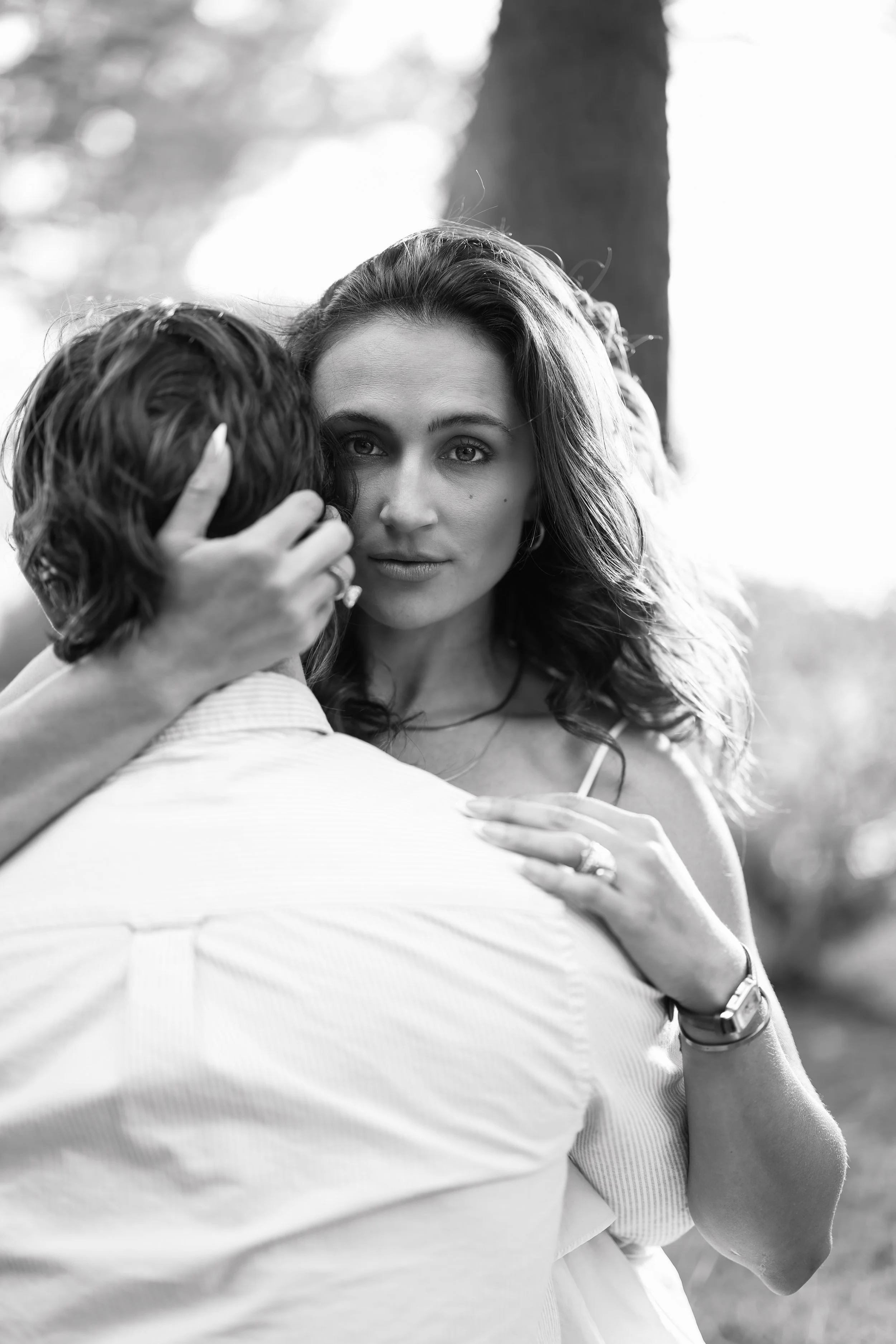 A black and white photo of a woman with wavy hair embracing a person whose face is not visible, against a backdrop of trees and sunlight.