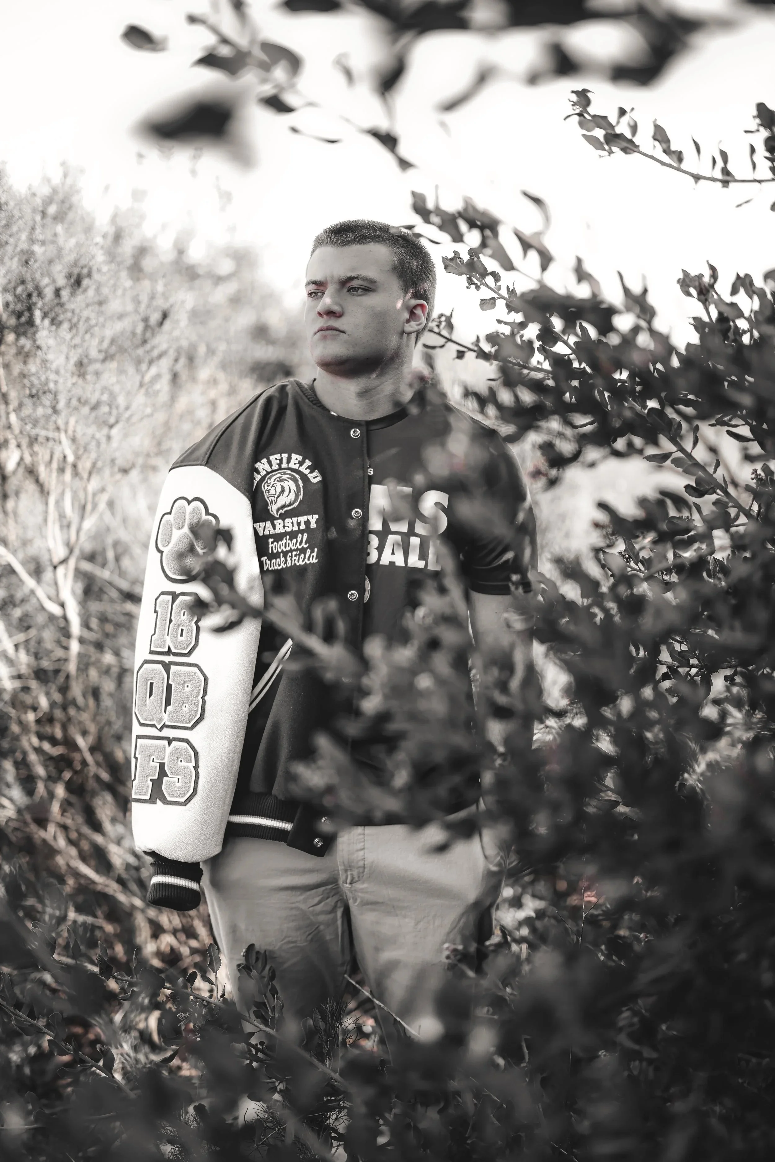A young man stands outdoors among bushes and trees, wearing a varsity jacket with patches and lettering, looking off into the distance with a serious expression.