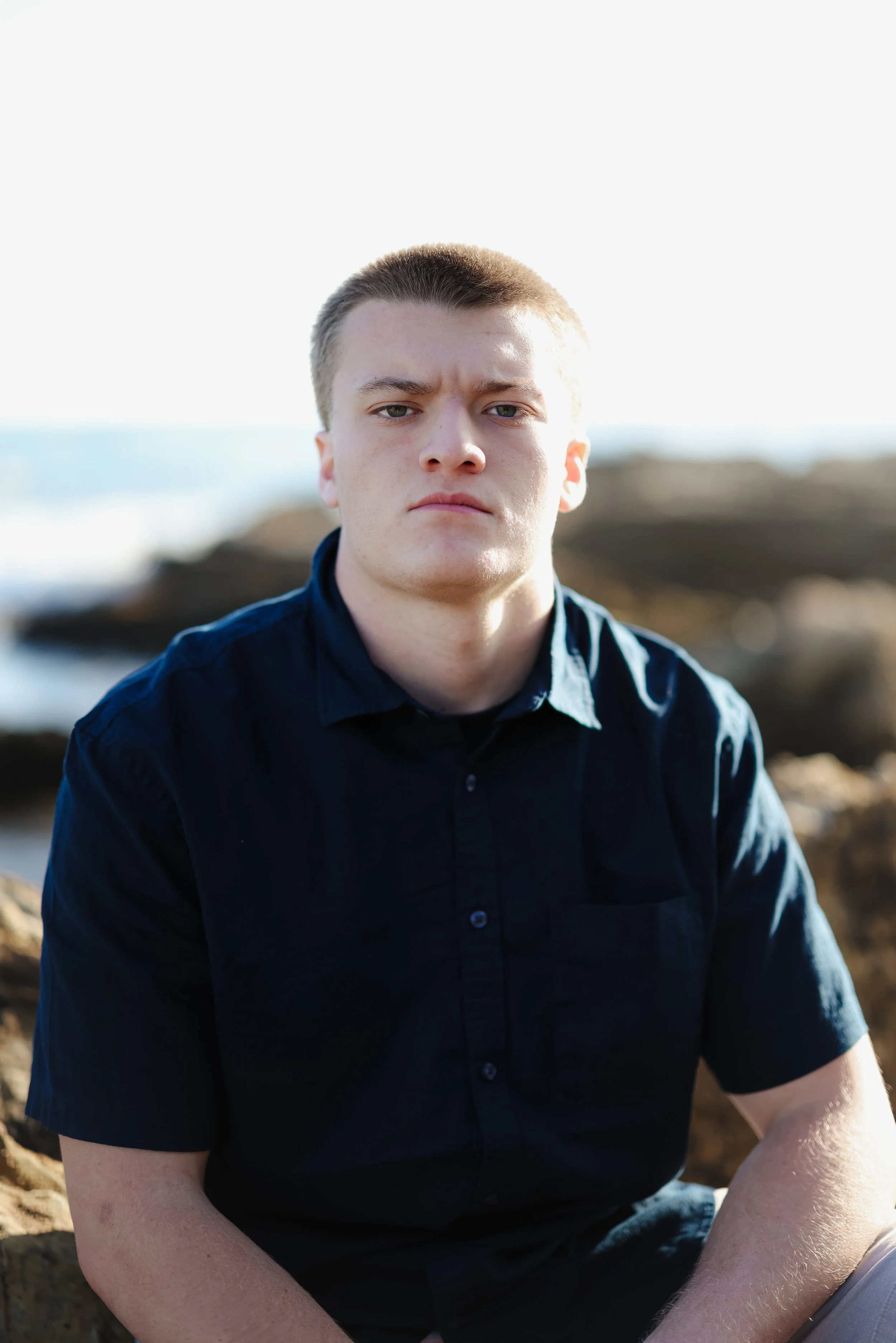 A young man with short light brown hair and fair skin sitting outdoors near rocks with the ocean in the background. He is wearing a dark blue button-up shirt and has a serious expression.