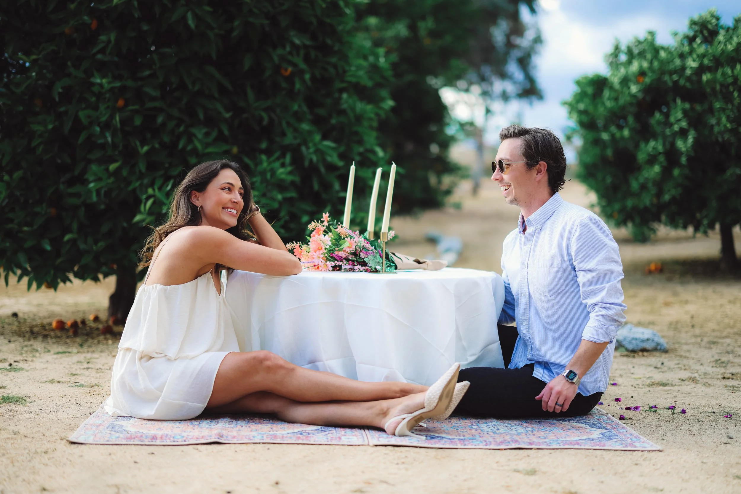A woman and a man enjoying a romantic moment outdoors at a picnic table with a floral centerpiece and candles. The woman is sitting on the ground with her legs extended, wearing a white dress and beige shoes, while the man, sitting cross-legged, is wearing a light blue shirt, sunglasses, and a smartwatch. They are smiling at each other, surrounded by green trees.