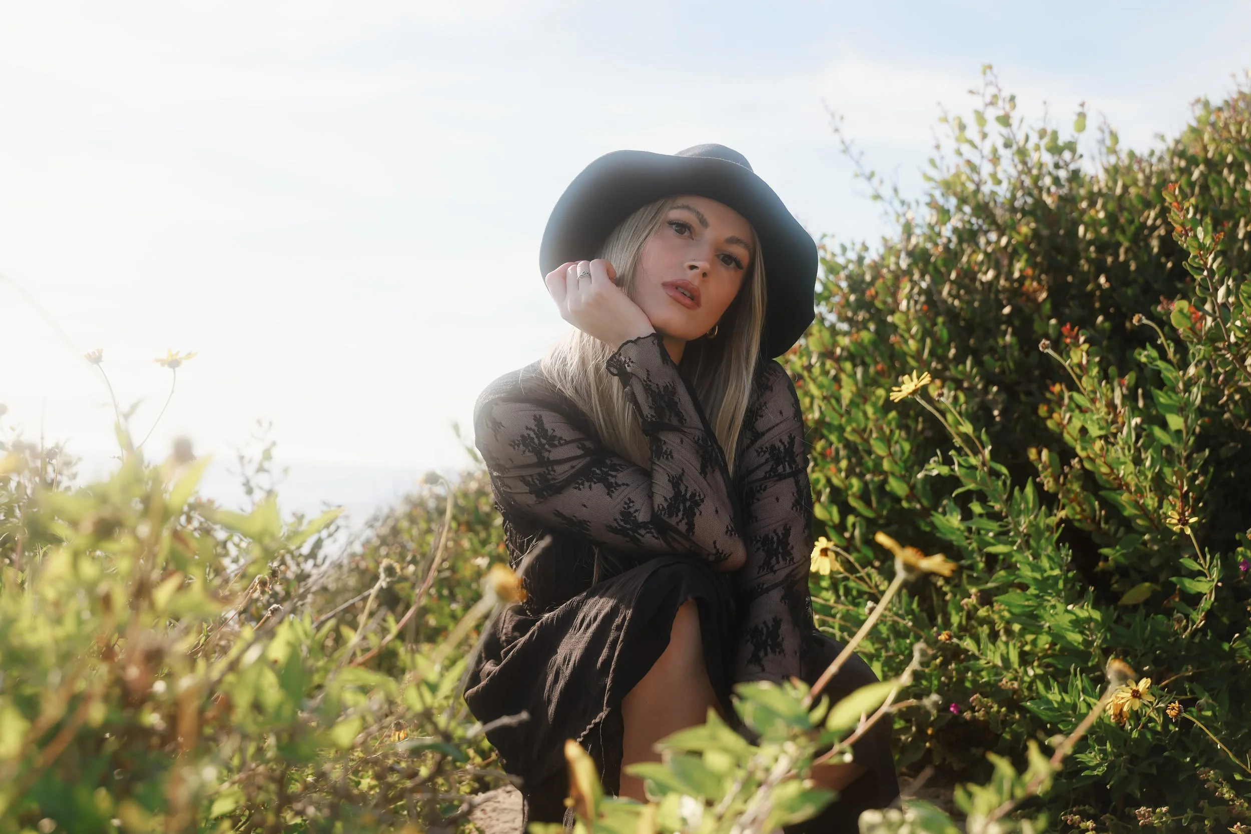 A woman in a black hat and black lace dress sitting among green bushes outdoors.