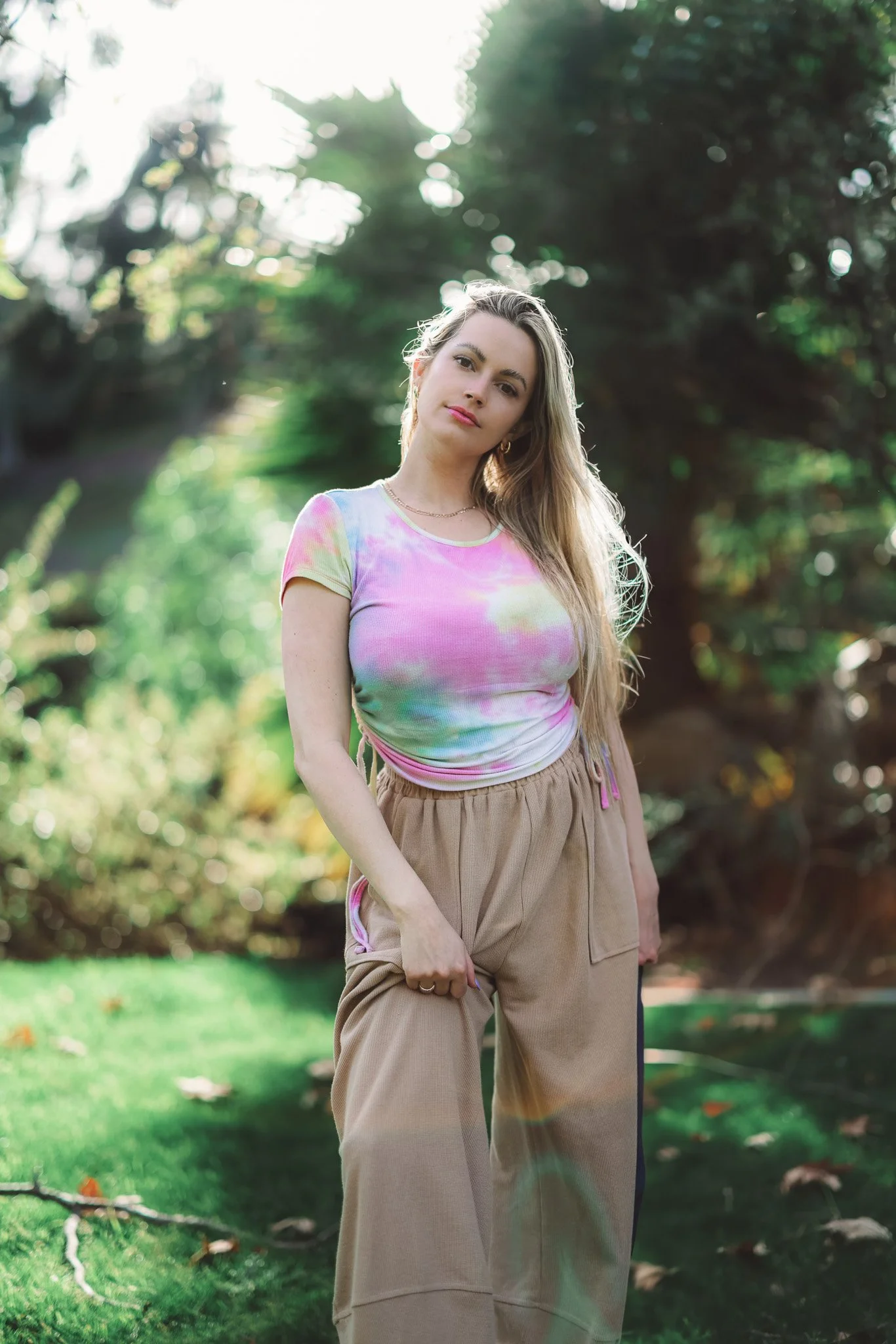 A young woman outdoors in a park-like setting with trees and greenery, wearing a tie-dye T-shirt and beige pants, looking at the camera with a neutral expression.