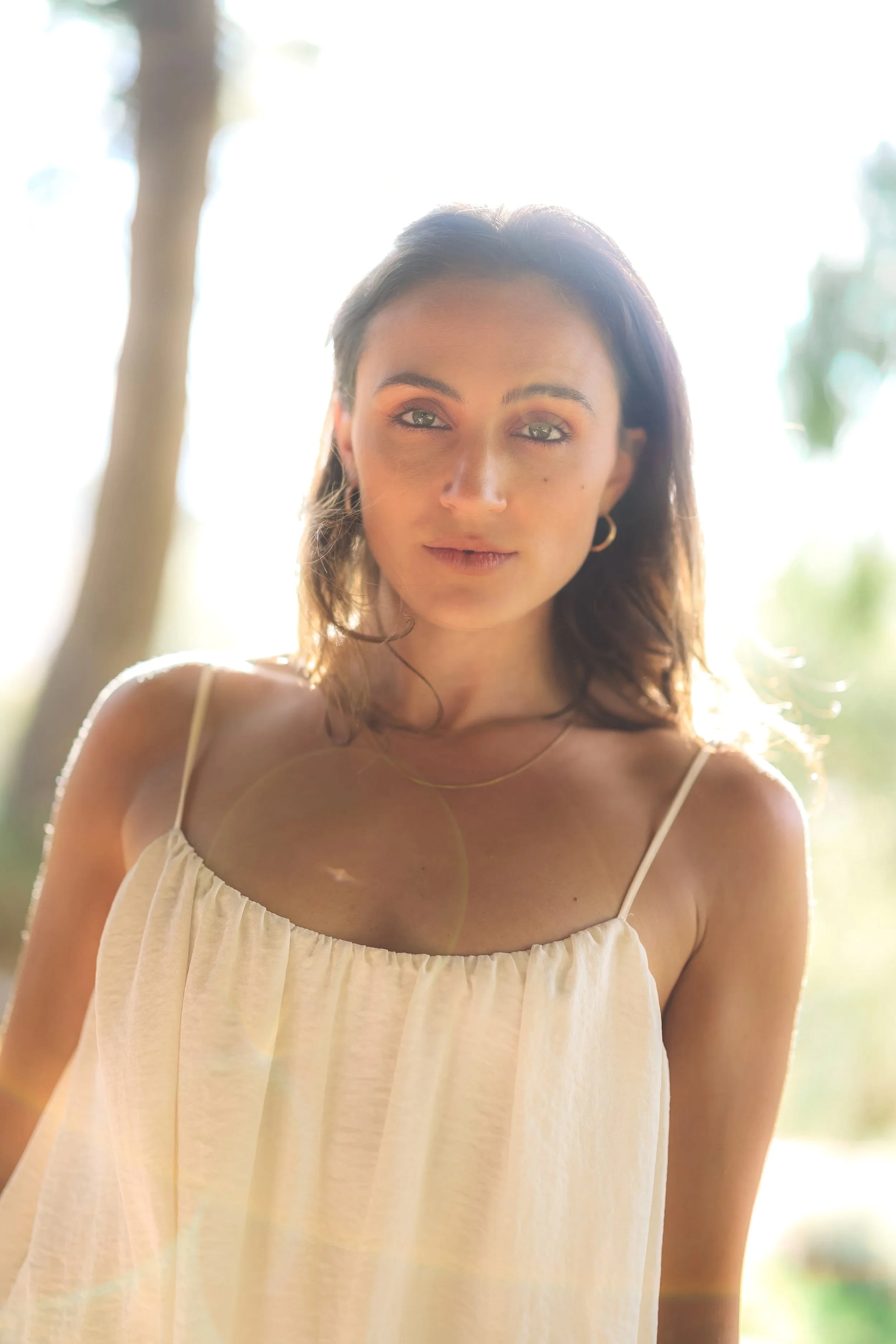 A young woman with brown hair and blue eyes standing outdoors with sunlight behind her, wearing a light-colored spaghetti strap dress and gold jewelry.