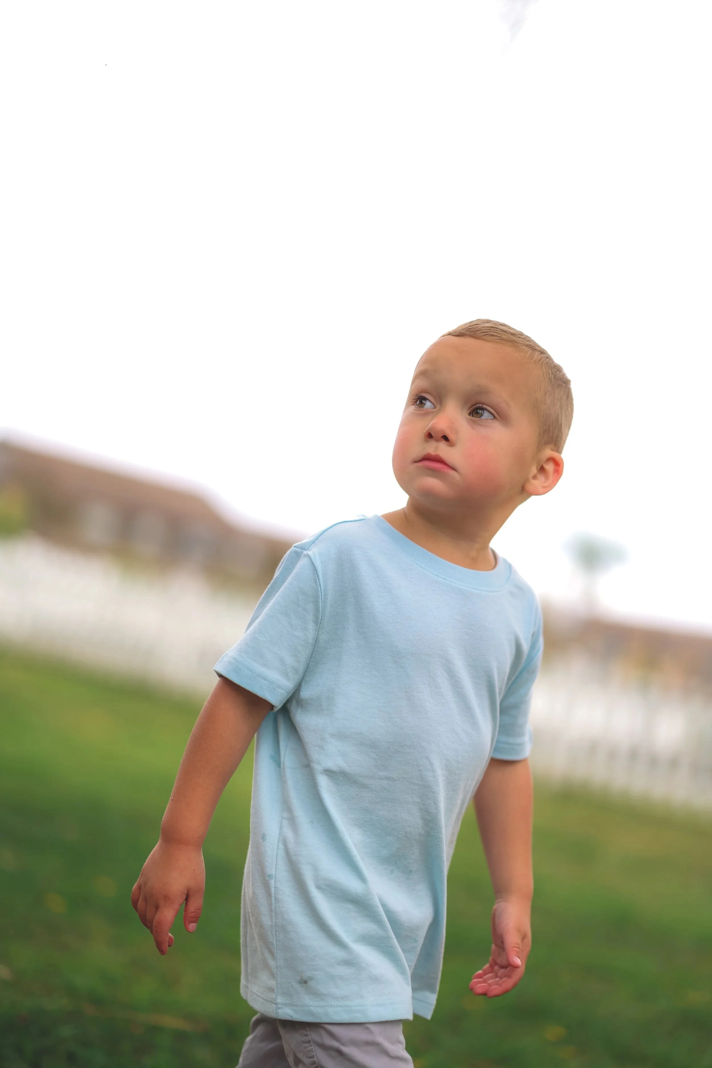 A young boy with short blond hair, wearing a light blue t-shirt and beige shorts, standing outdoors on grass, looking to the right with a serious expression.