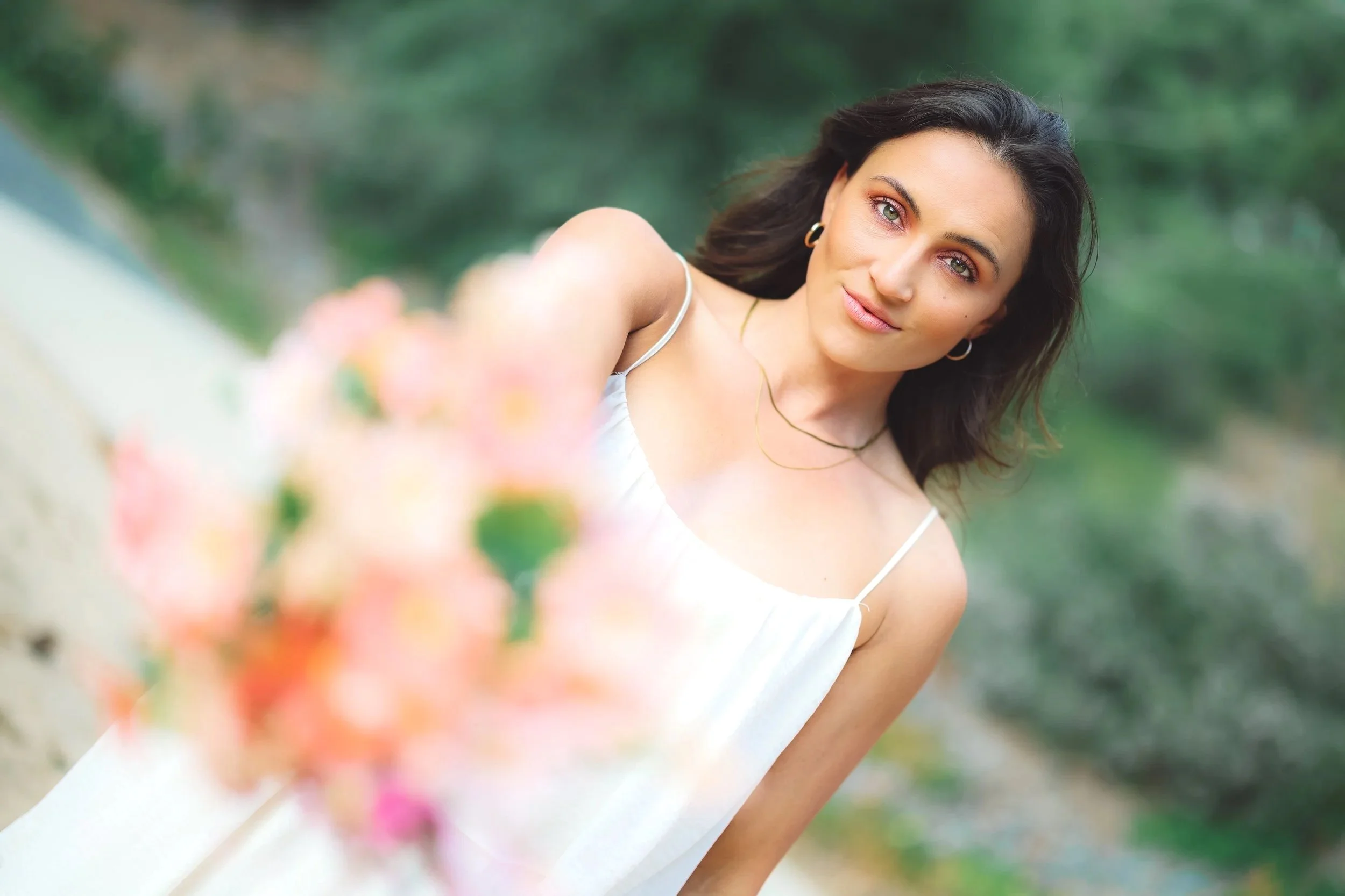 A young woman with dark hair and green eyes standing outdoors, wearing a white dress and gold jewelry, holding a bouquet of pink flowers slightly out of focus in the foreground.