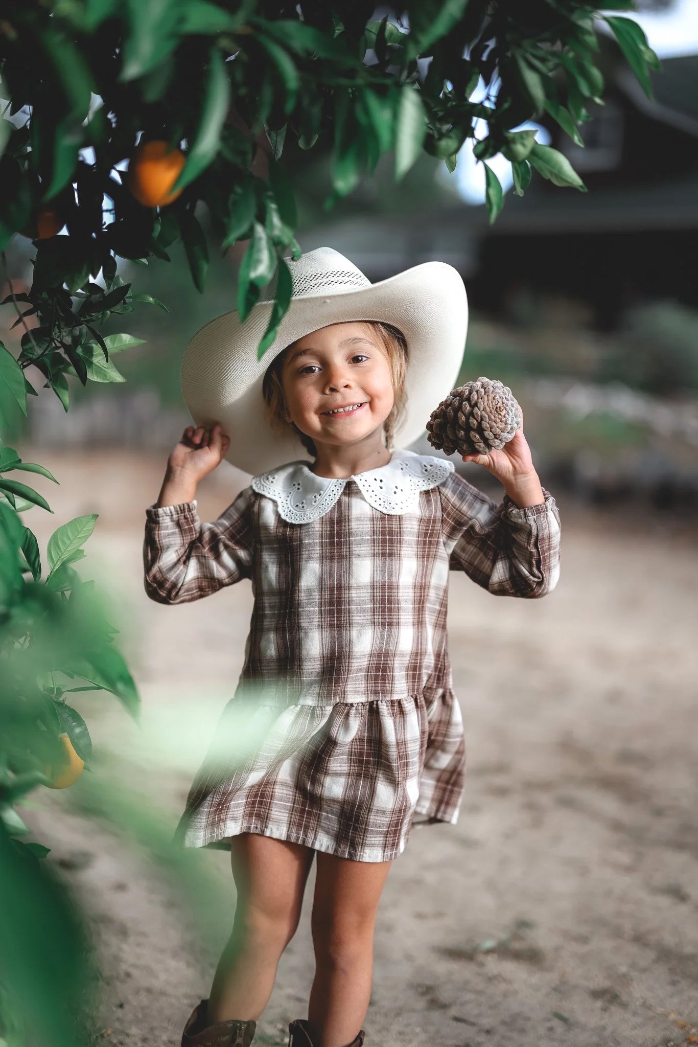 A young girl wearing a plaid dress, cowboy boots, and a large white hat, holding a pine cone and smiling in an orchard.