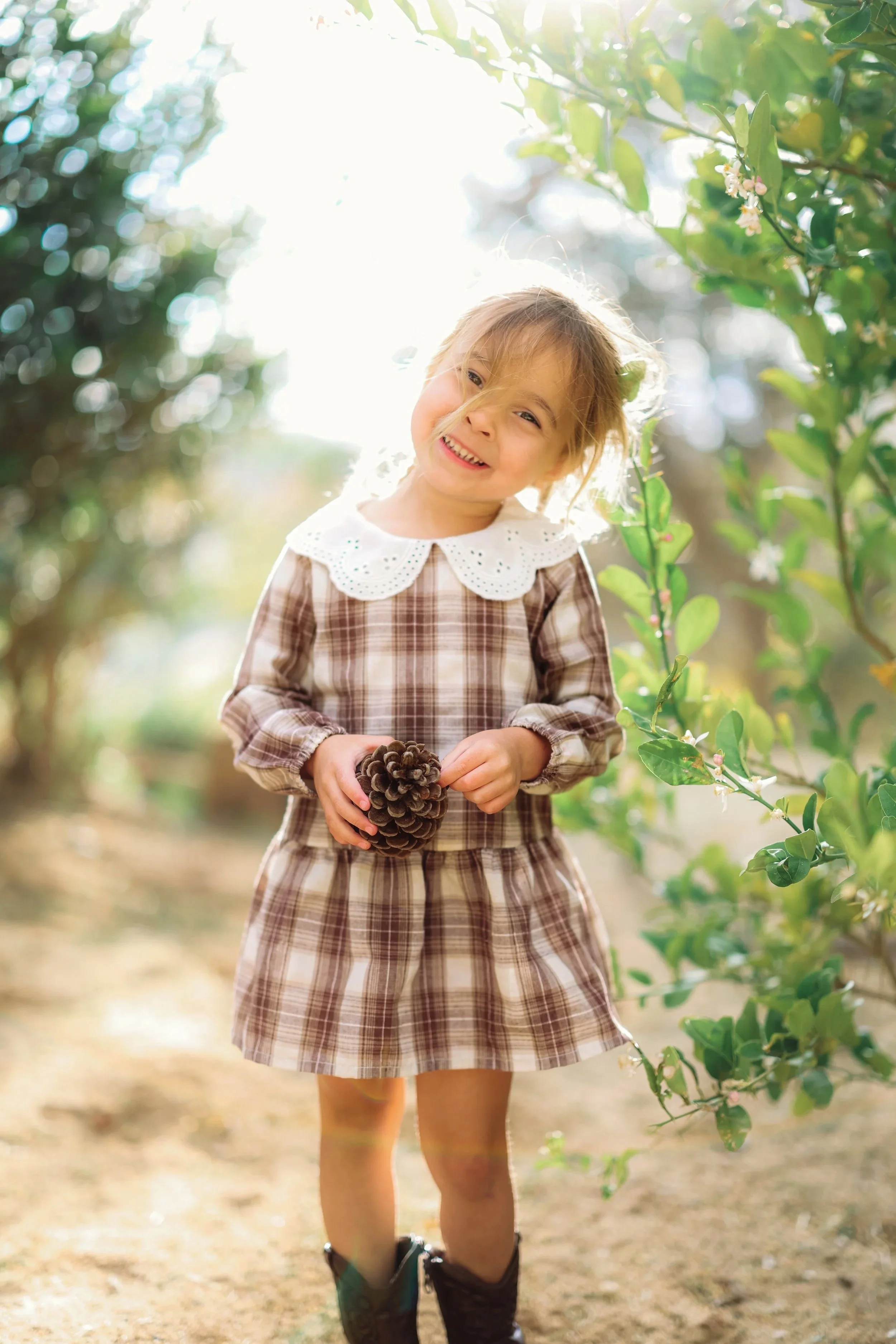 A young girl smiling and holding a pine cone in her hands, standing outdoors among green plants and sunlight.