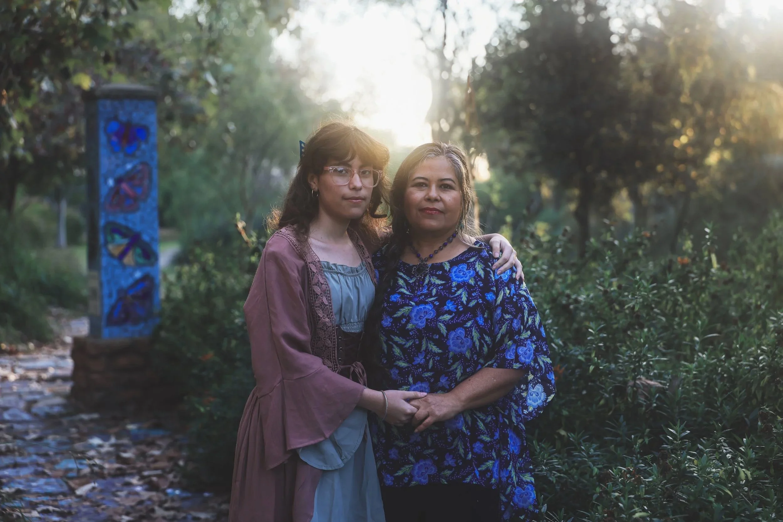 Two women standing closely together outdoors at sunset, with trees and a blue painted sculpture in the background.