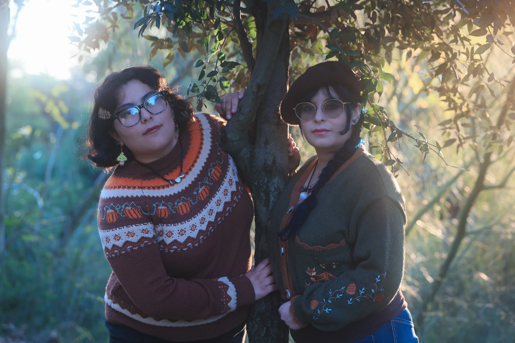 Two women standing outdoors near a tree, both wearing glasses and sweaters with holiday or winter-themed patterns, with sunlight filtering through the branches.