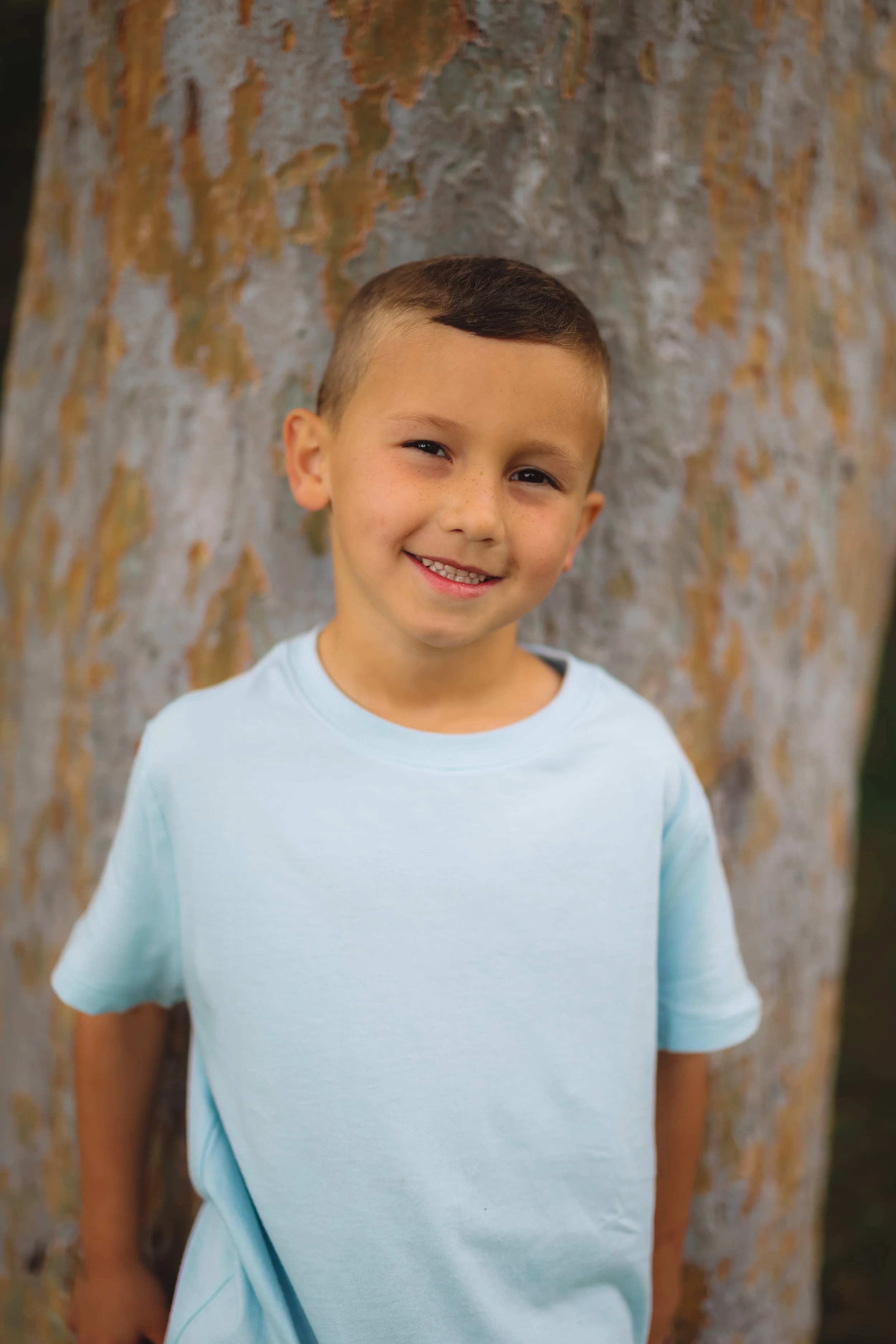 Young boy smiling, standing in front of a large tree trunk with colorful bark.
