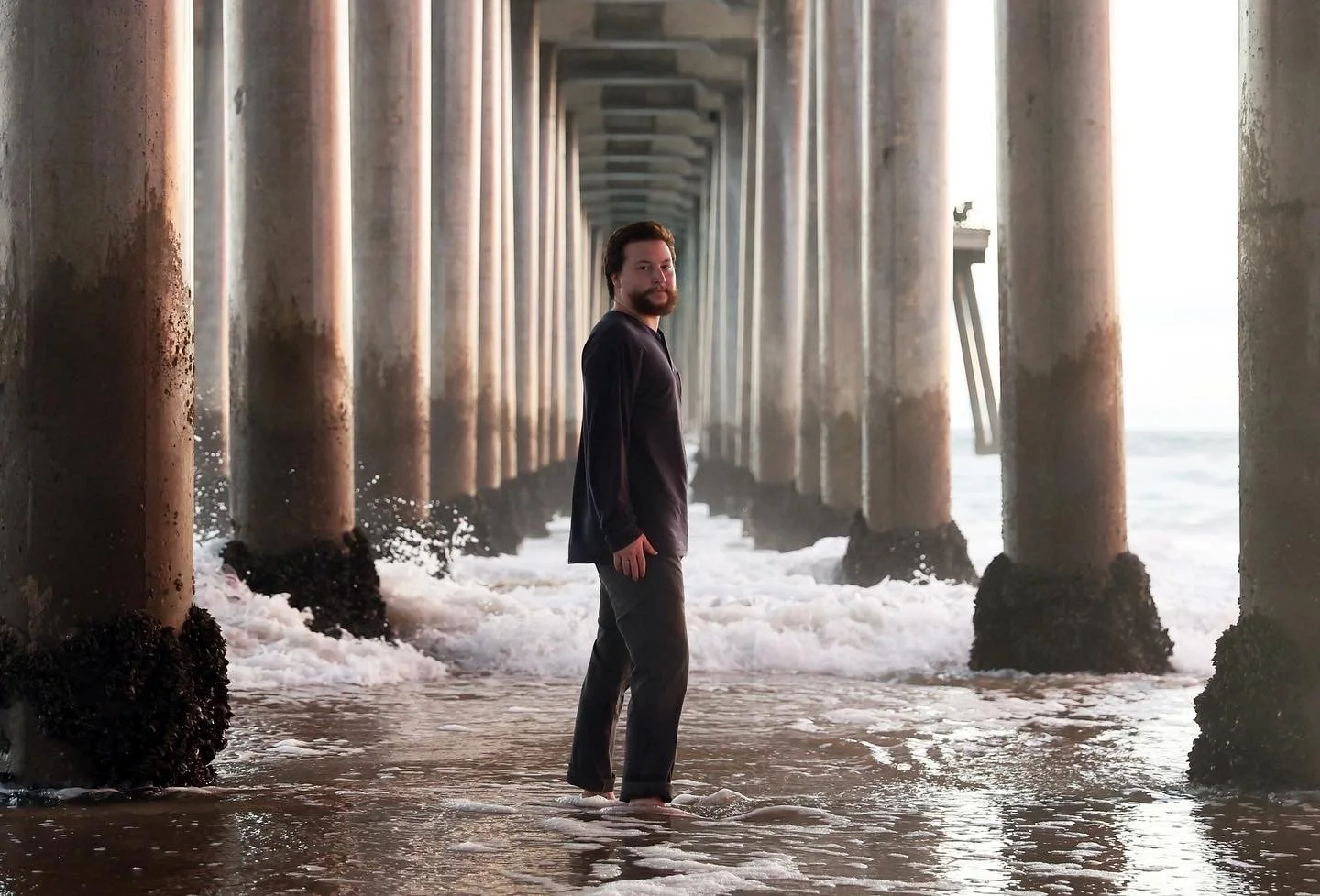 A man standing in the shallow water under a pier with concrete support columns, waves around his feet, looking at the camera, with the ocean in the background.