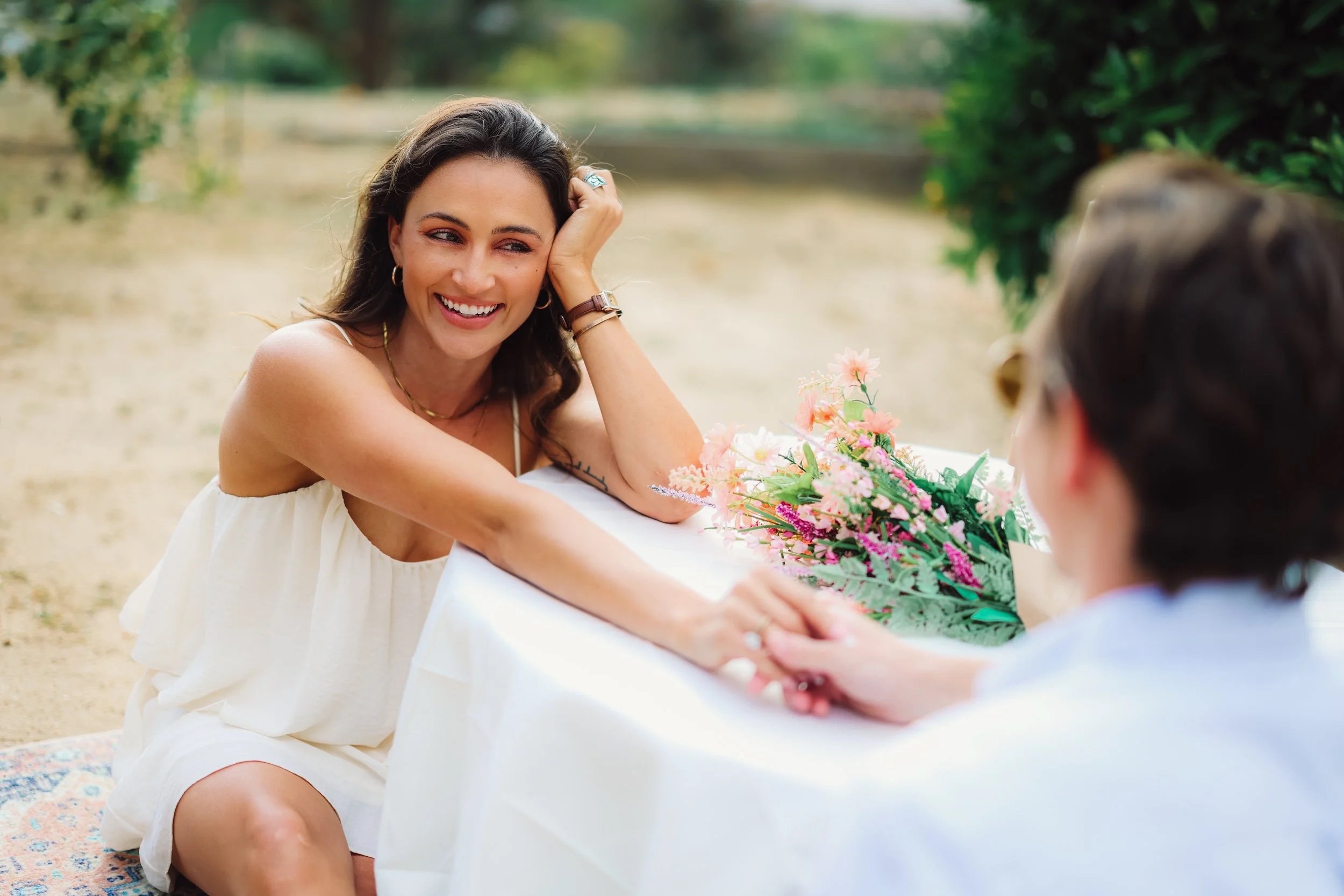 A woman in a white dress smiling and touching her head, sitting at a table with a bouquet of pink flowers, outdoors in a lush green setting.
