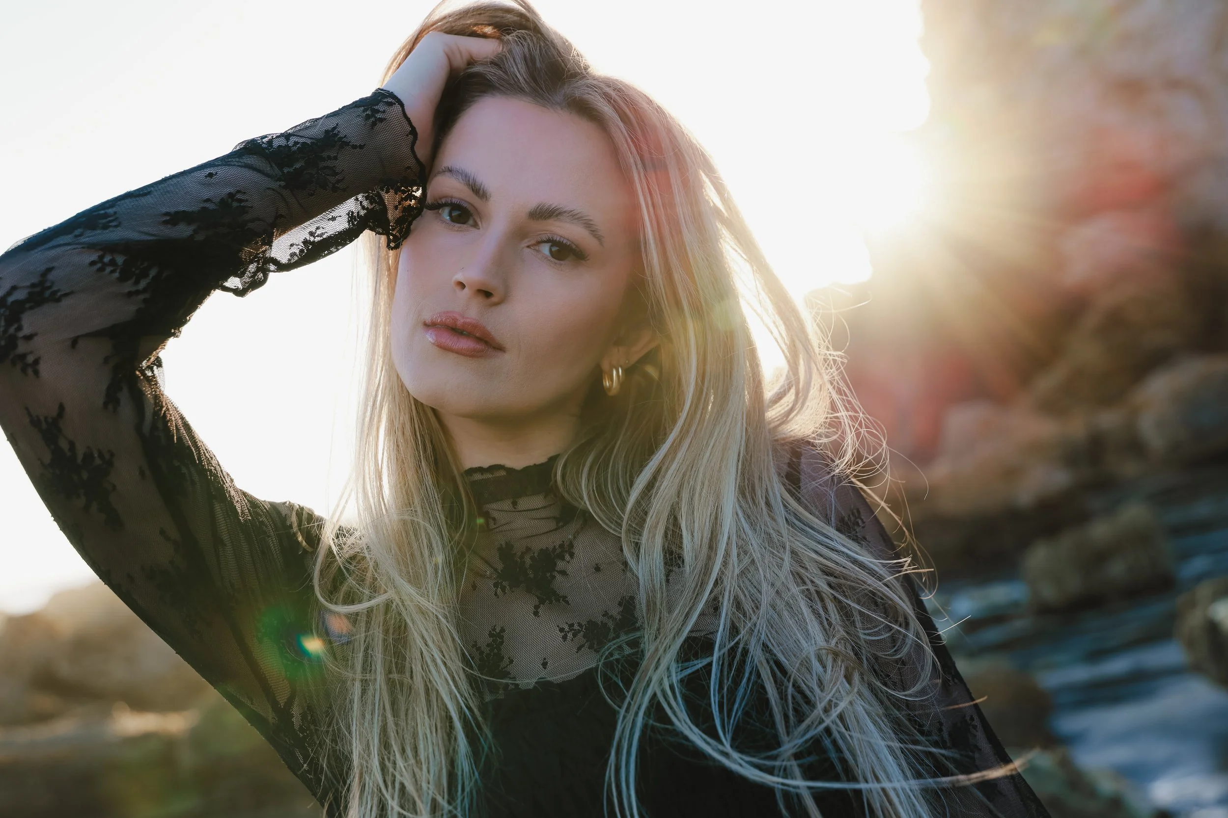 A young woman with long blonde hair wearing a black lace top, standing outdoors at sunset with the sun glowing behind her.