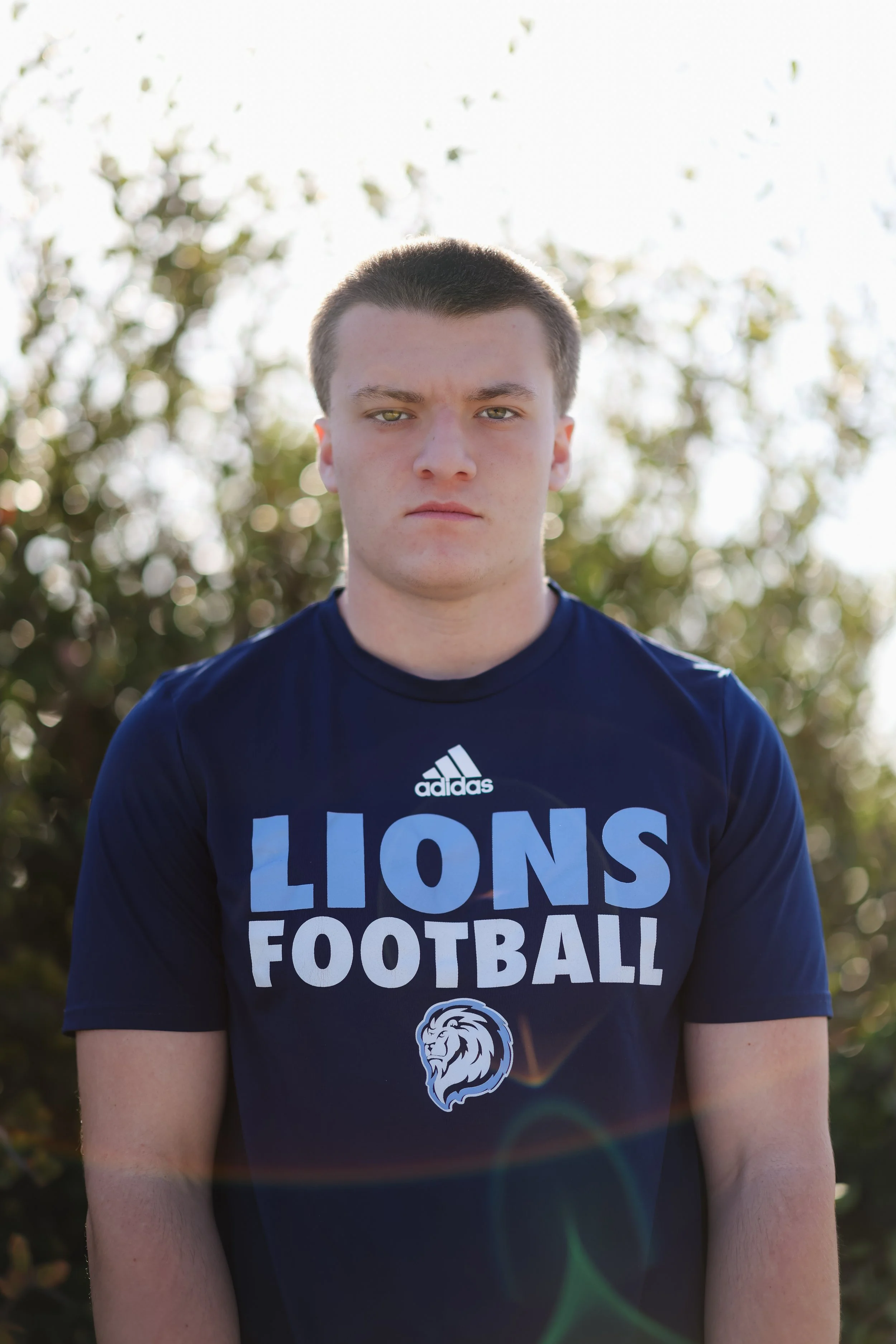 Young man wearing a navy blue Lions Football shirt, standing outdoors with plants behind him, facing the camera with a serious expression.