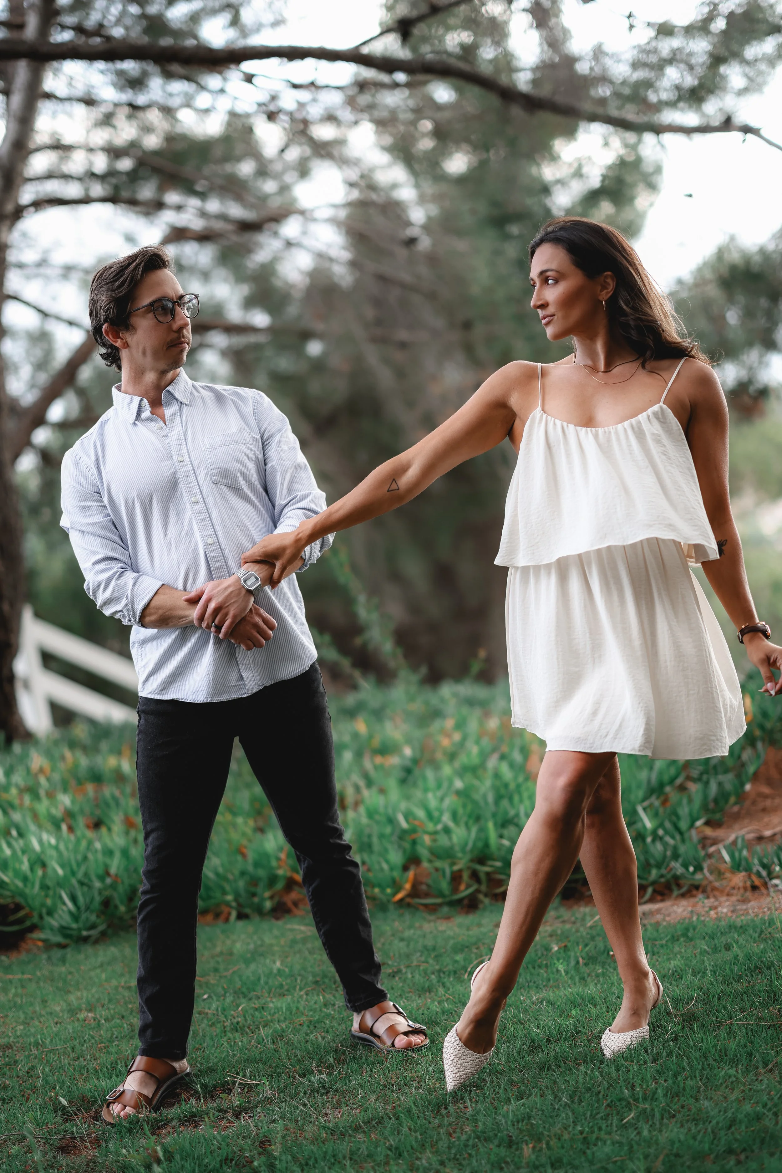 A woman in a white sundress is pulling a man's arm, who is standing nearby on a grassy area with trees in the background. The man is wearing glasses, a light-colored button-up shirt, black pants, and sandals.
