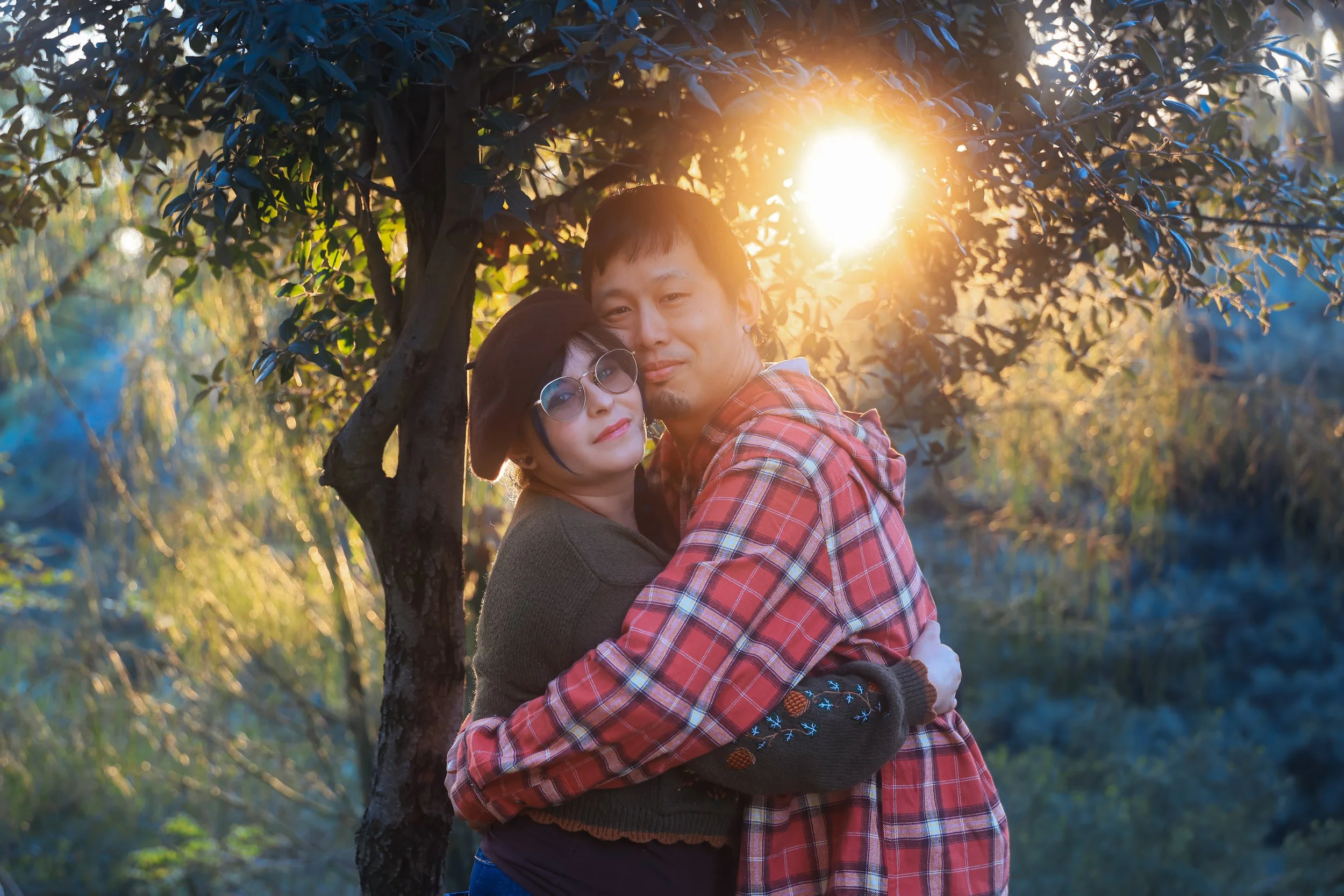 A couple hugging in a park at sunset, with the sun shining through the trees in the background.