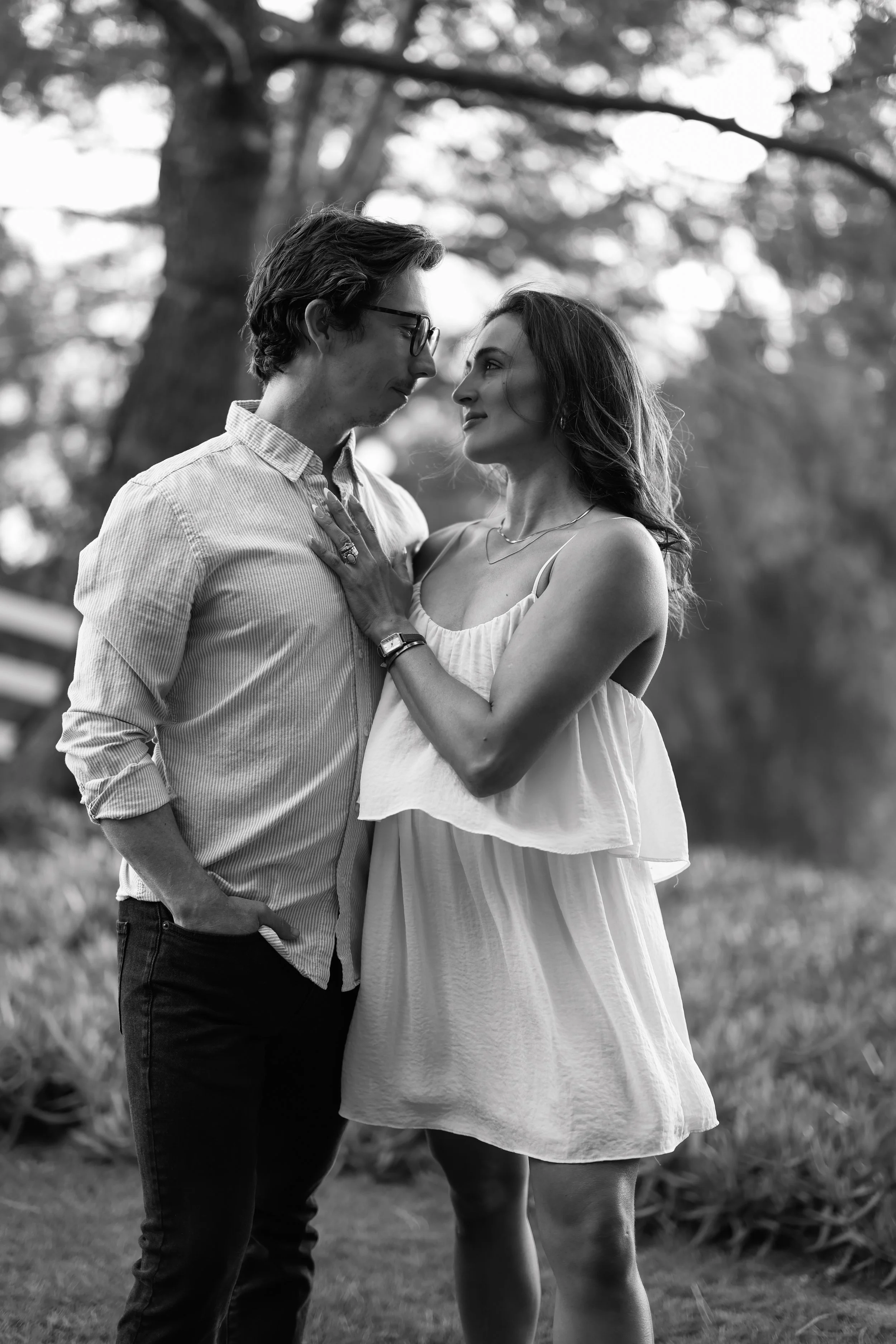 A black-and-white photo of a man and woman facing each other outdoors under a tree, with the woman touching the man's chest and looking into his eyes.