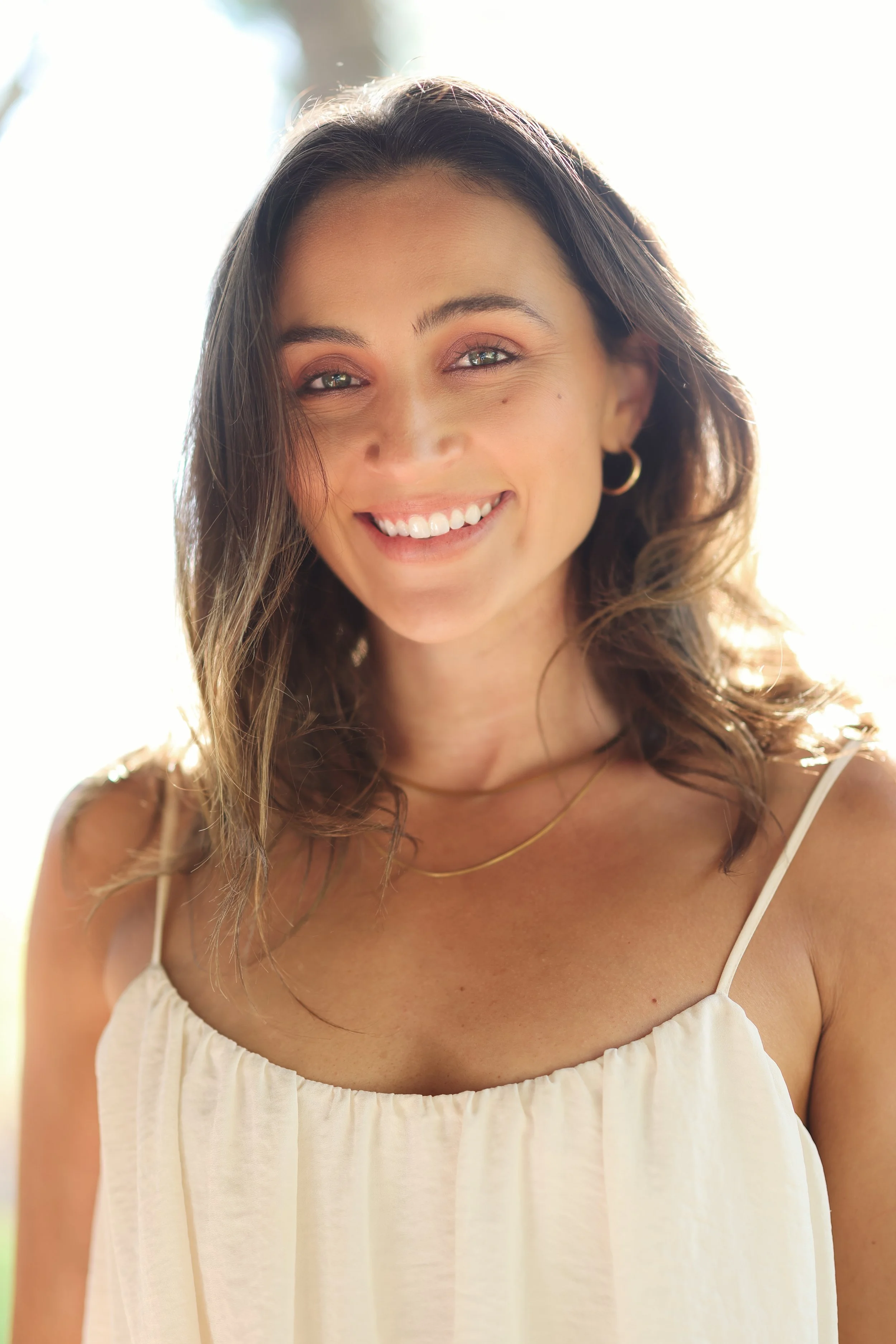 A woman smiling outdoors in bright sunlight, wearing a white spaghetti strap top and gold jewelry.