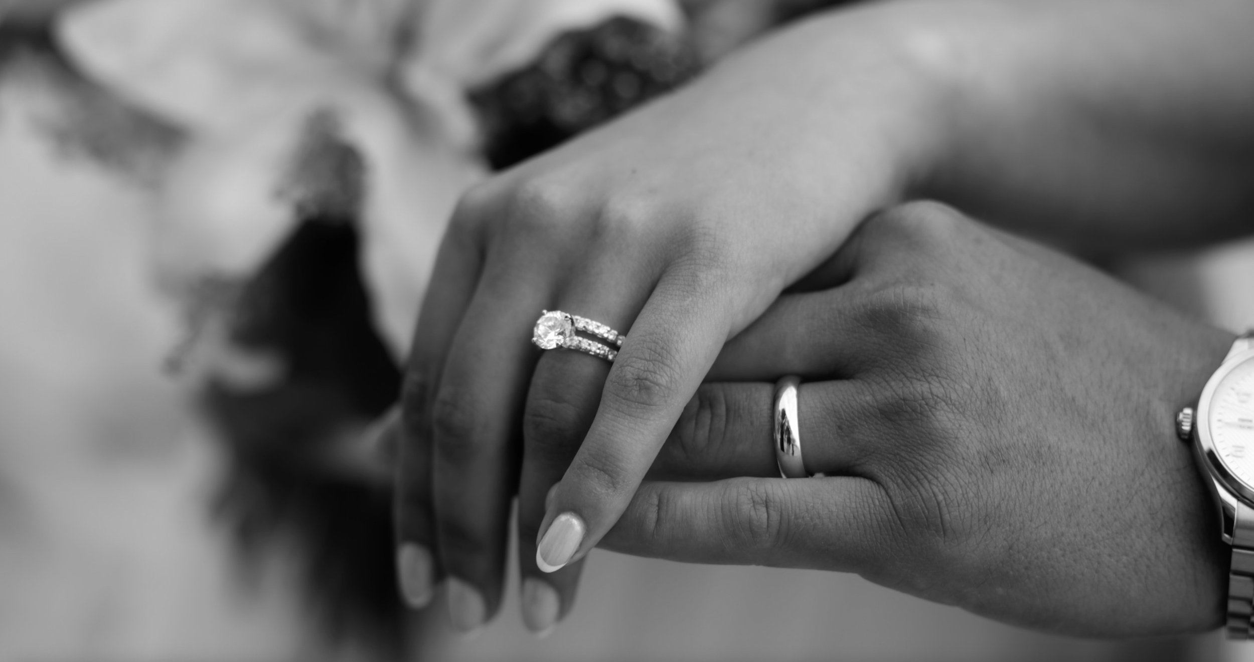 Close-up of a couple's hands holding each other, with the woman's hand displaying a diamond engagement ring and a wedding band, and the man's hand wearing a wedding band and a watch, in black and white.