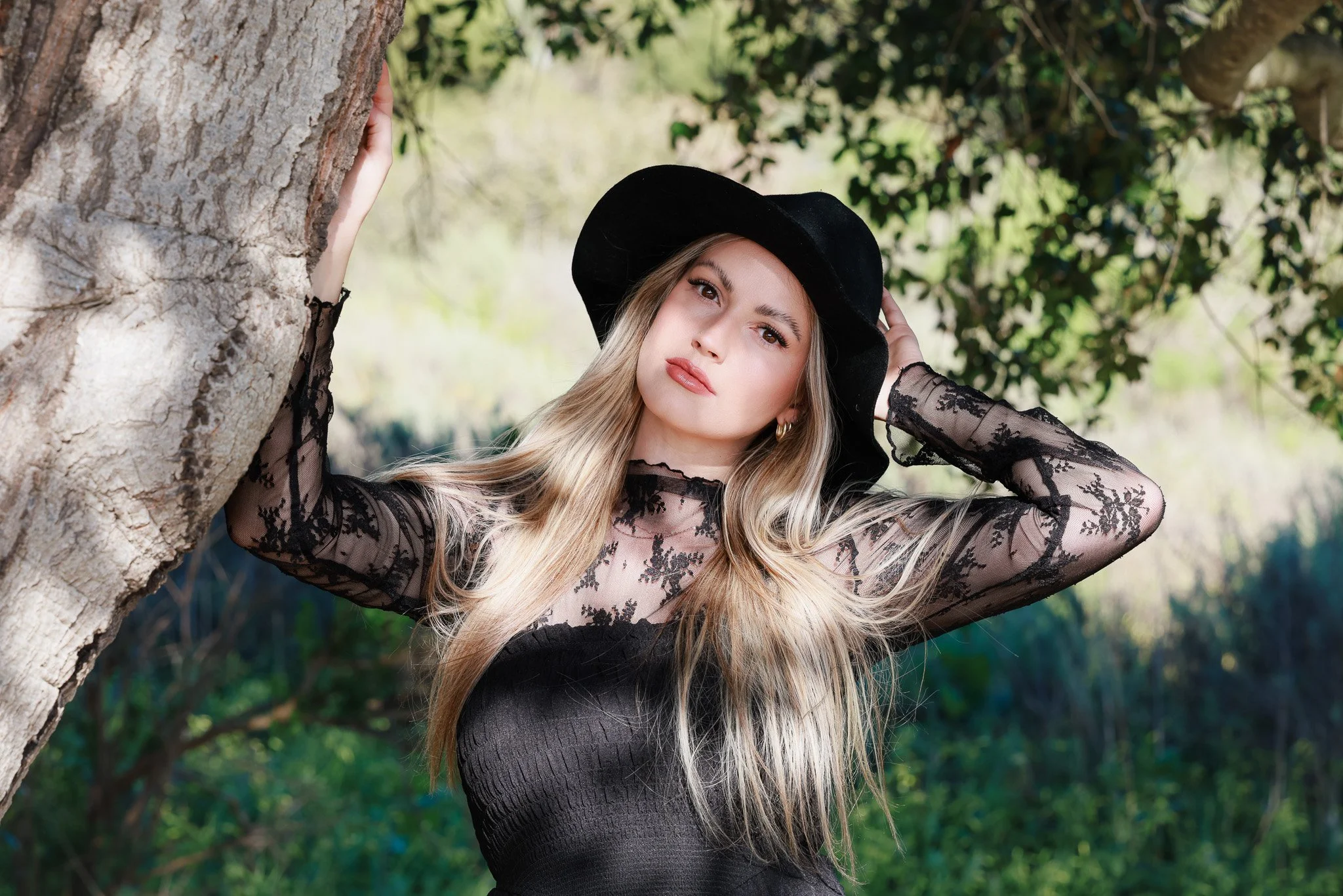A woman with long blonde hair wearing a black lace dress and a wide-brimmed black hat, leaning against a tree outdoors with greenery in the background.