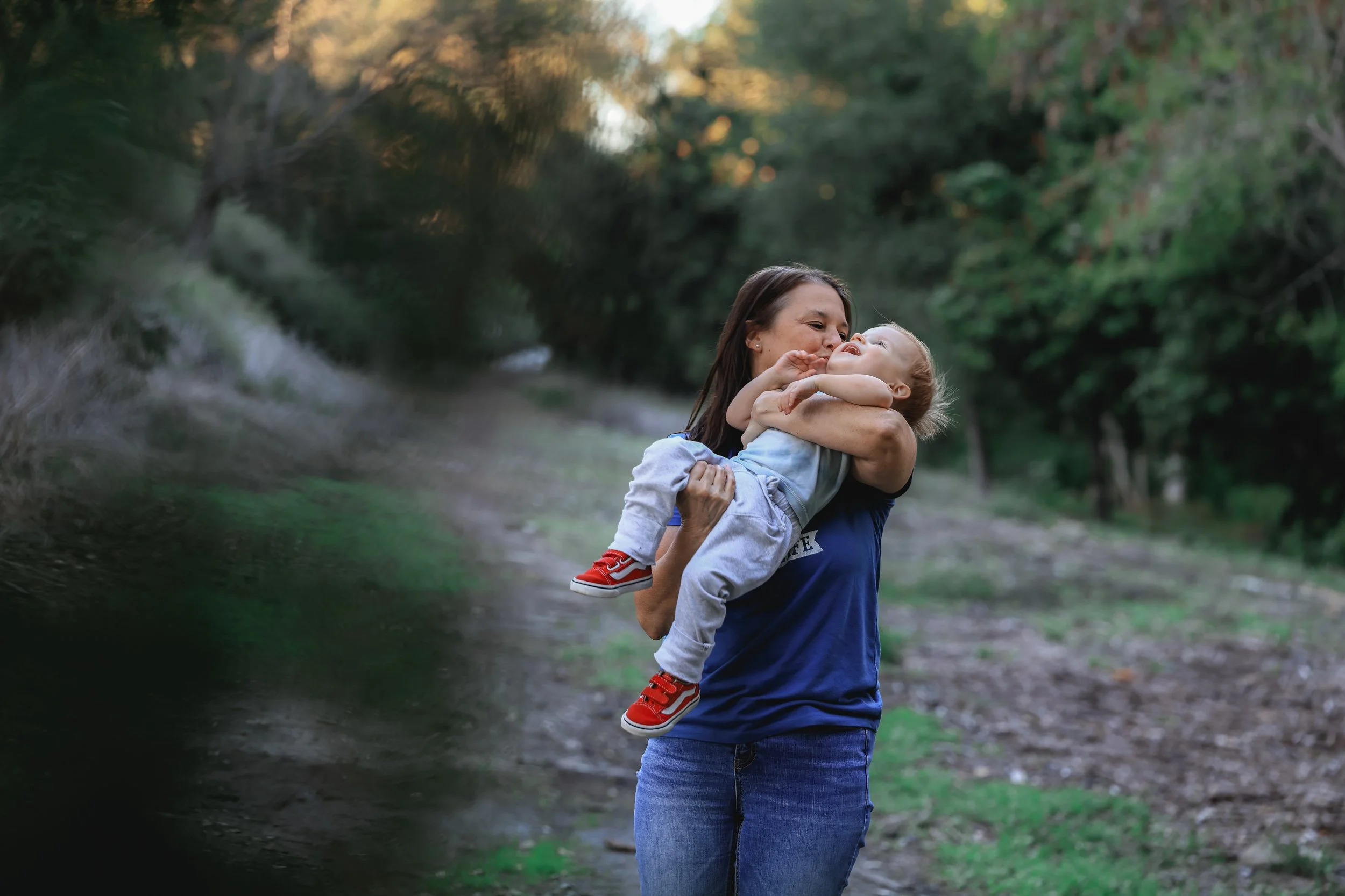 A woman holding a young child in her arms outdoors on a dirt path surrounded by trees, with the woman smiling and the child reaching up with both hands.