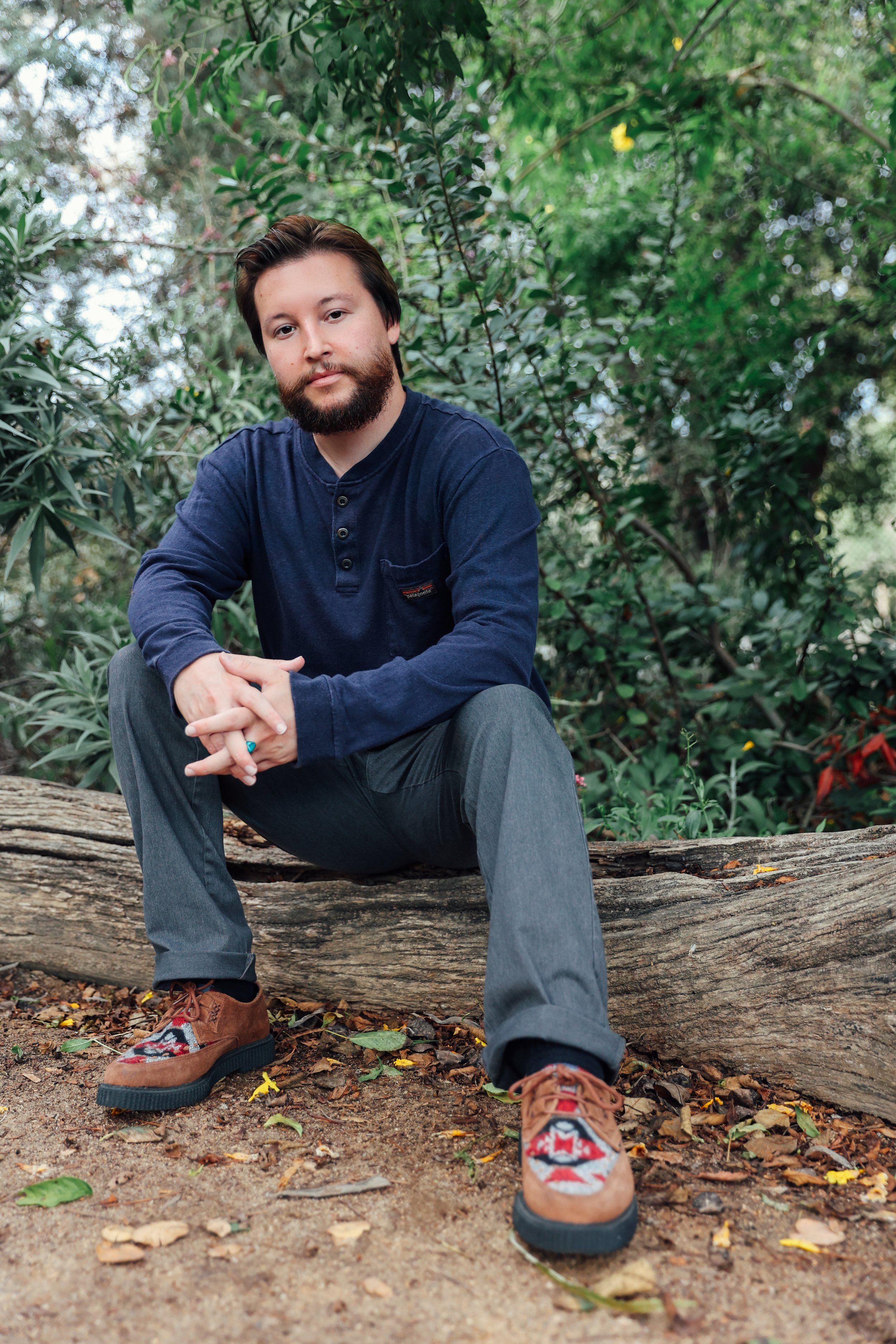 A man with a beard and dark hair sitting on a fallen tree trunk outdoors in a lush, green forest. He is wearing a navy blue long-sleeve shirt, gray pants, and colorful patterned shoes.