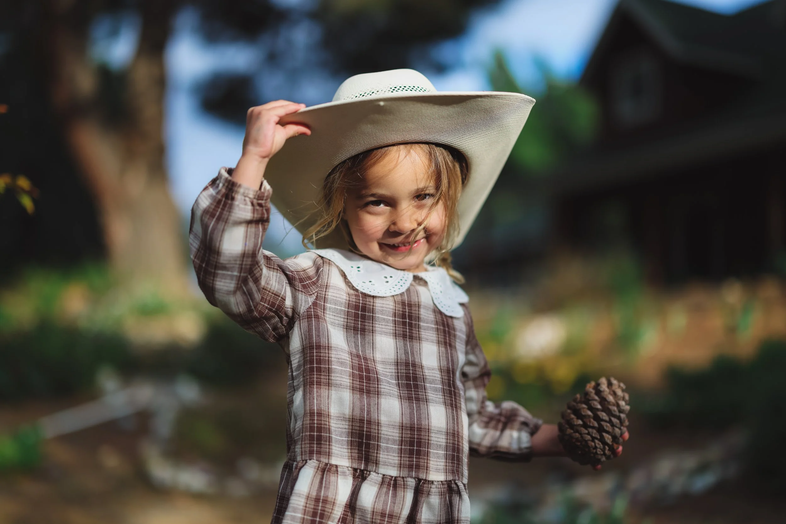 A young girl with a big smile, wearing a plaid dress and white sunhat, holding a pinecone in her hand outdoors.