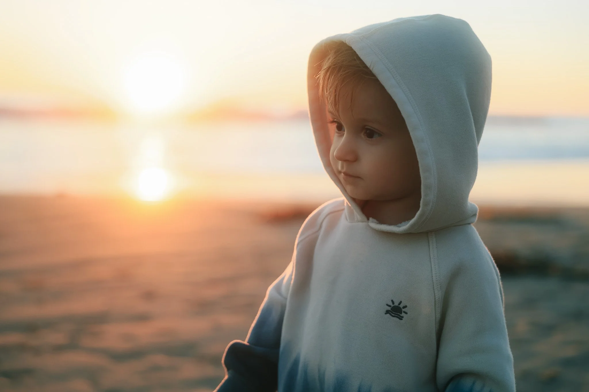 A young boy in a white hoodie standing on the beach during sunset, looking thoughtfully at the horizon.